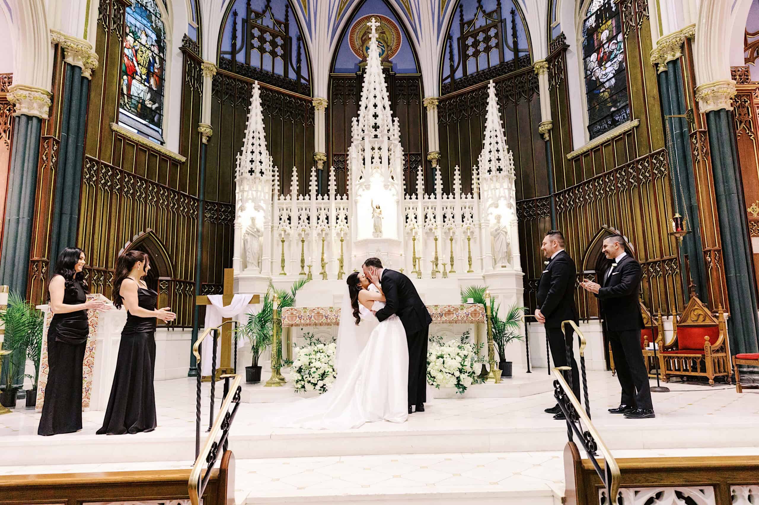 A bride and groom kiss at the altar during a Classic Wedding at Ballroom at the Ben, with bridesmaids and groomsmen standing on either side in a beautifully decorated church.