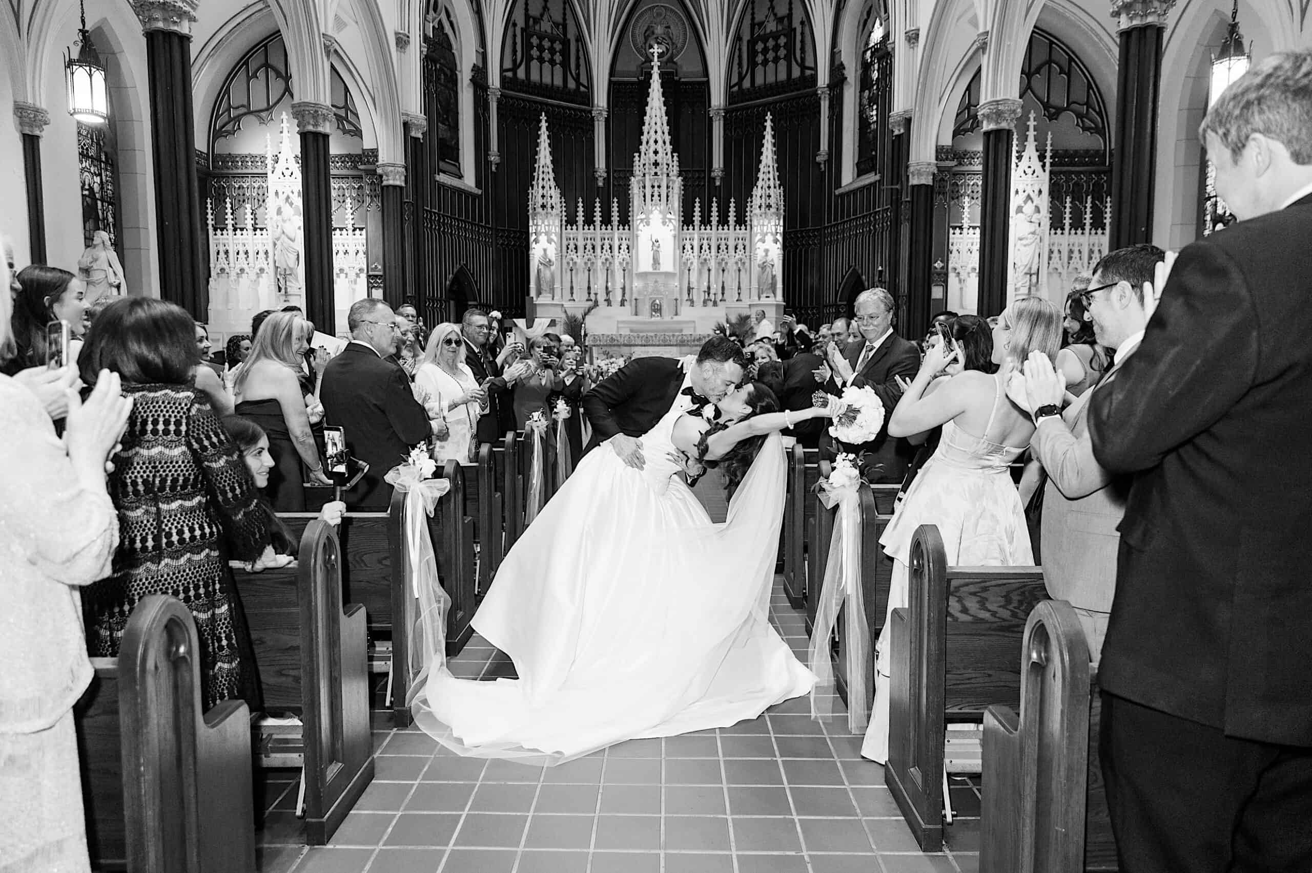 A bride and groom kiss at the end of the aisle in a church as guests watch and applaud during their Classic Wedding at Ballroom at the Ben.