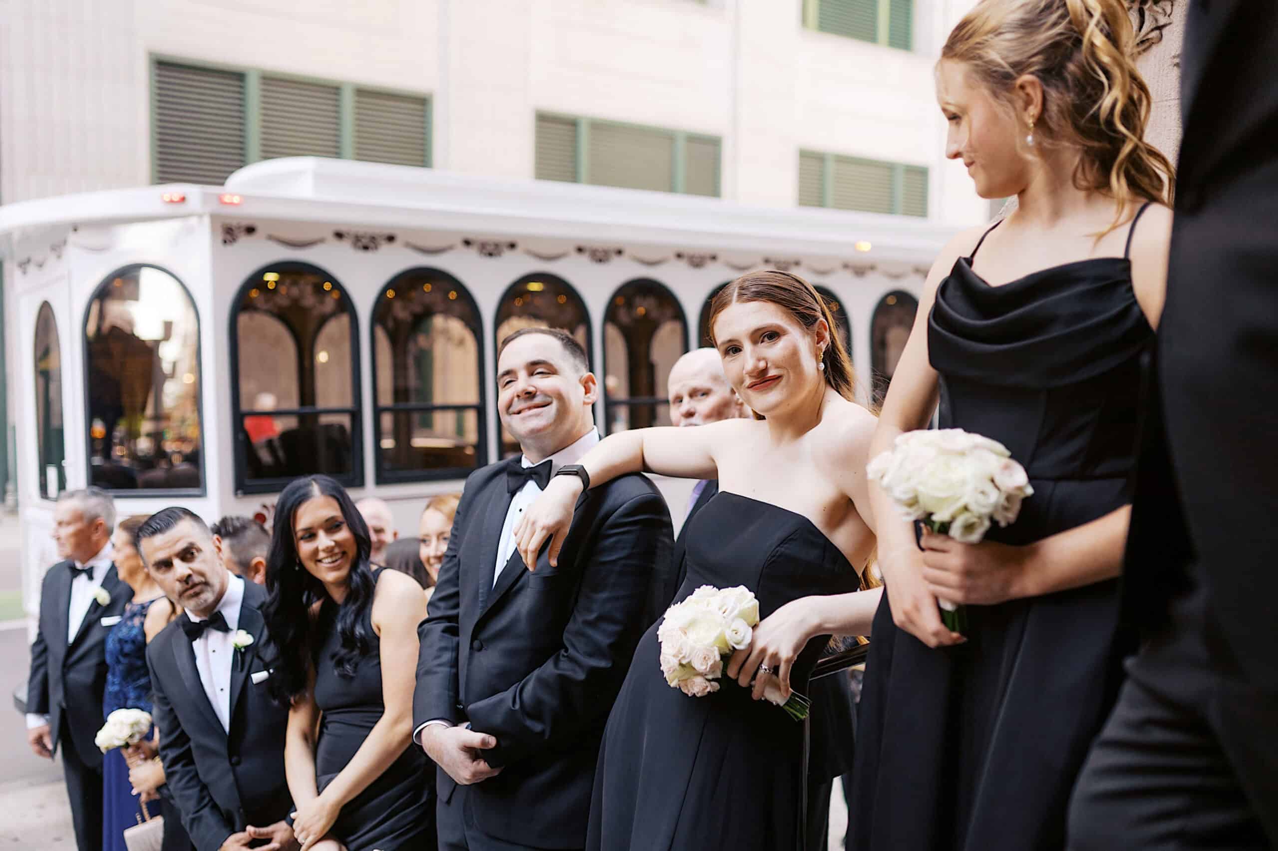 A group of people dressed in formal black attire stand in a line outdoors, some holding white bouquets, with a white trolley bus in the background—capturing the elegance of a Classic Wedding at Ballroom at the Ben.
