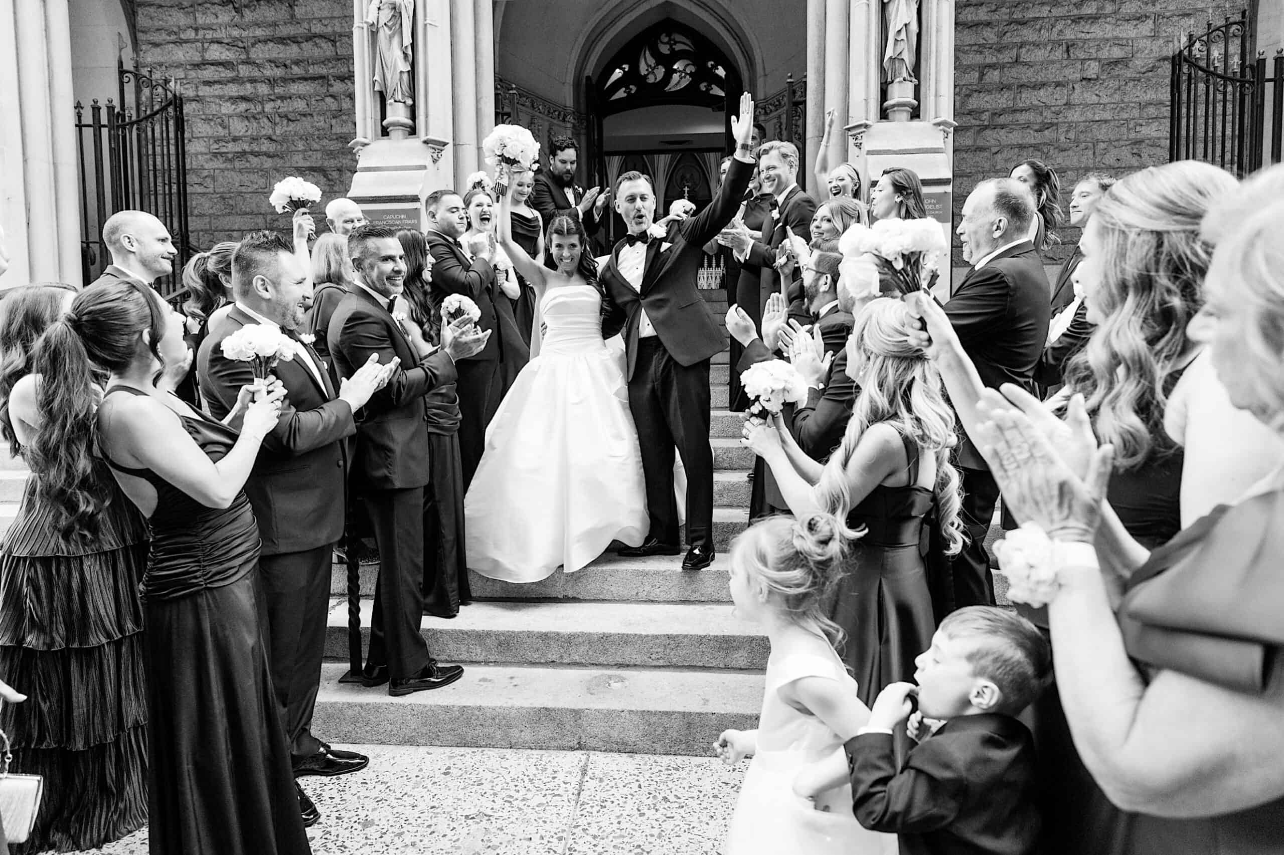 A bride and groom celebrate on church steps, surrounded by applauding guests dressed formally; flower bouquets and smiling faces fill the scene in this Classic Wedding at Ballroom at the Ben.