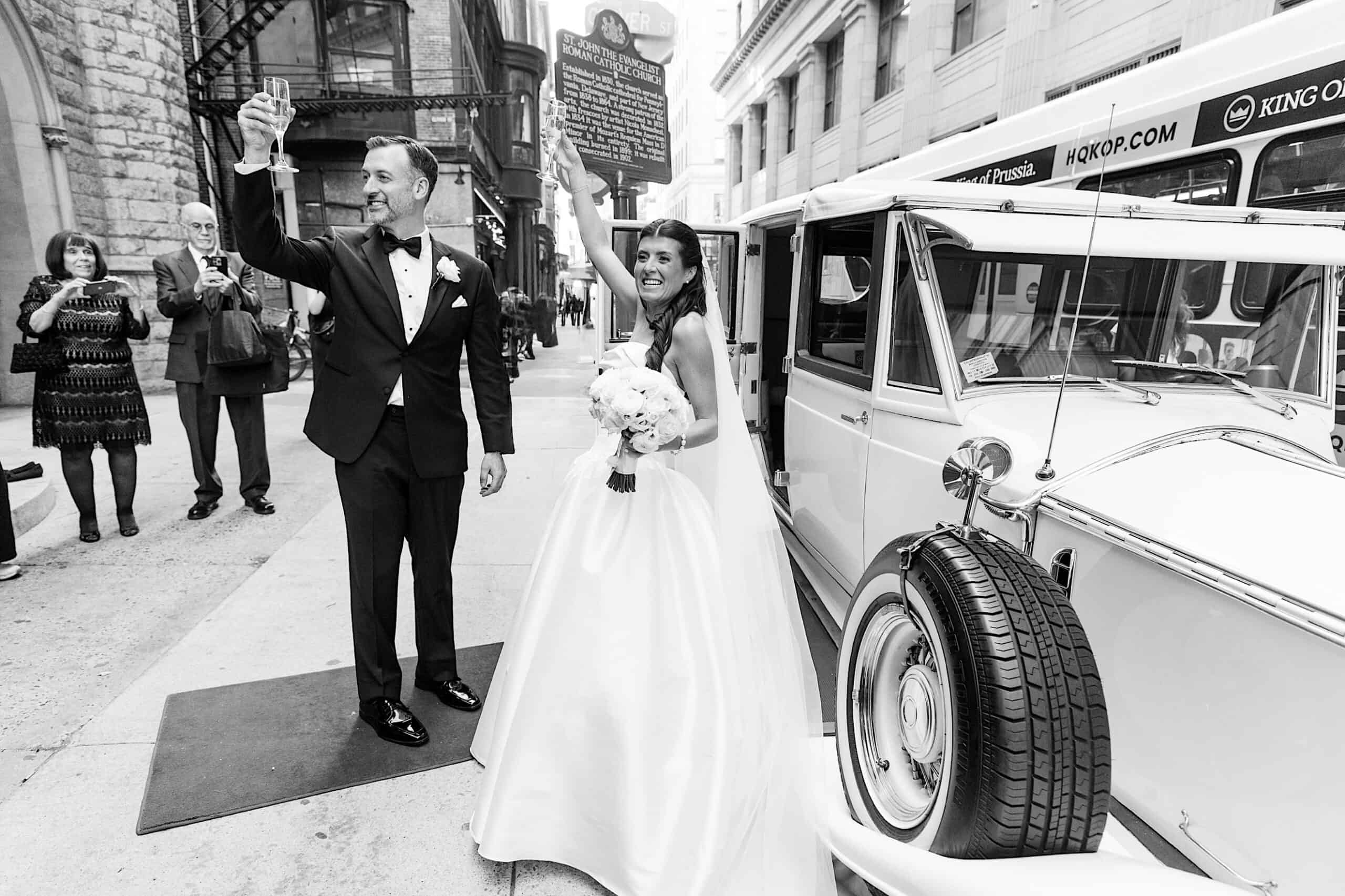 A bride and groom stand beside a vintage car on a city street; the groom raises a glass in a toast while the bride smiles, capturing the elegance of their Classic Wedding at Ballroom at the Ben.