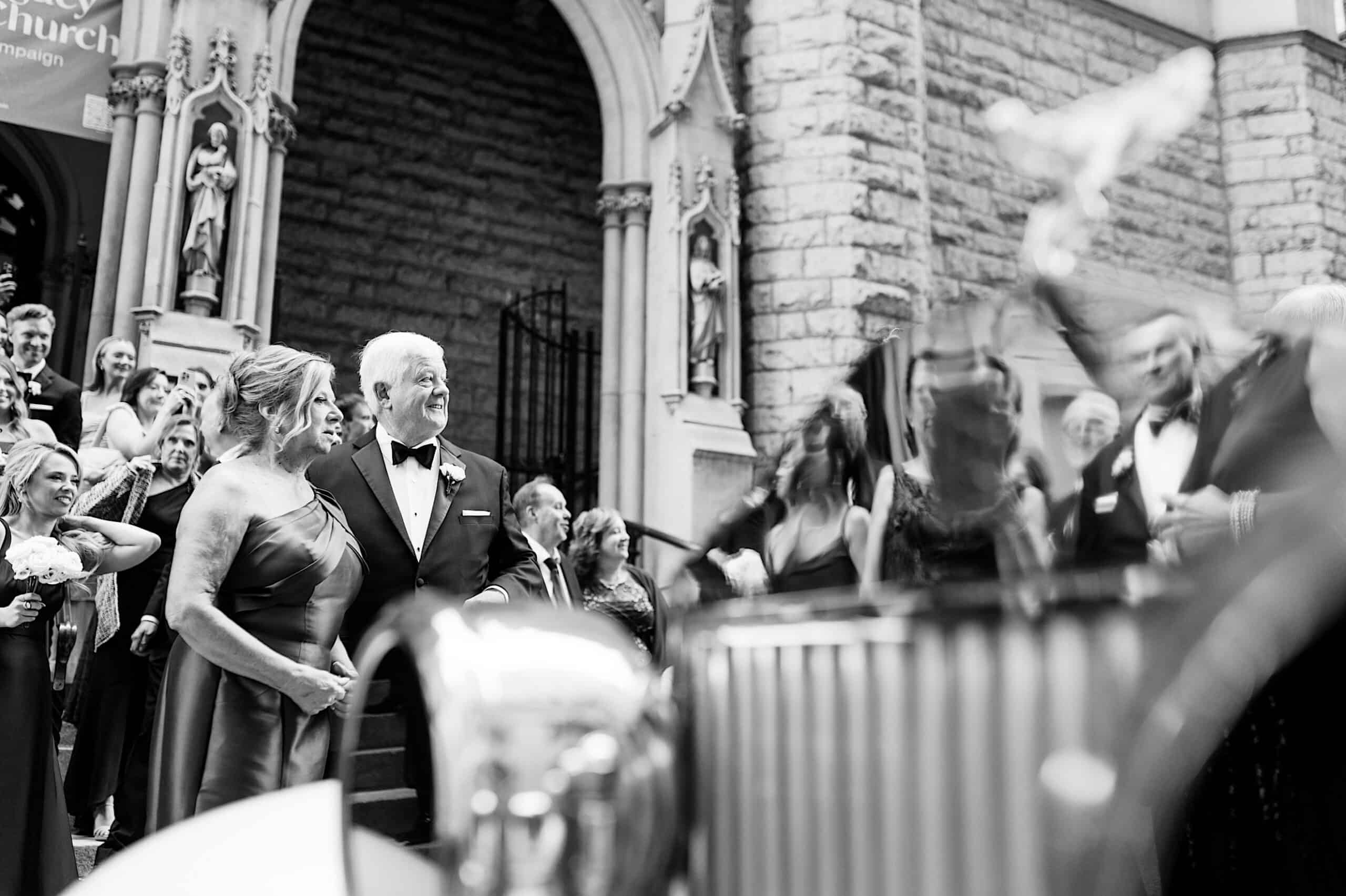 A well-dressed couple stands smiling on church steps, surrounded by guests in formal attire, capturing the elegance of a Classic Wedding at Ballroom at the Ben. The background features stone architecture and statues, with a slightly blurred foreground.