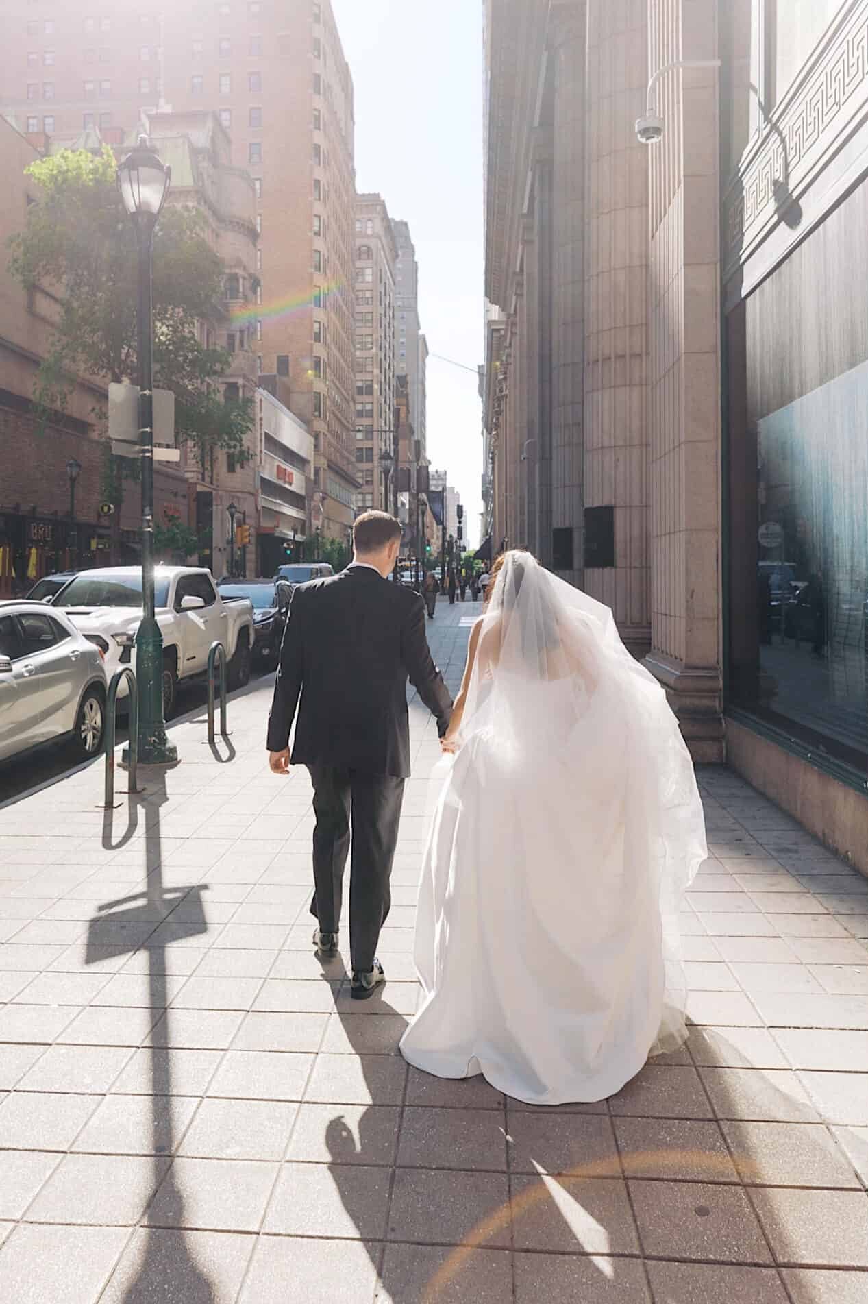 A bride and groom in wedding attire walk through a city street and pose together in front of a historic building with a clock tower, capturing timeless moments from their Classic Wedding at Ballroom at the Ben.