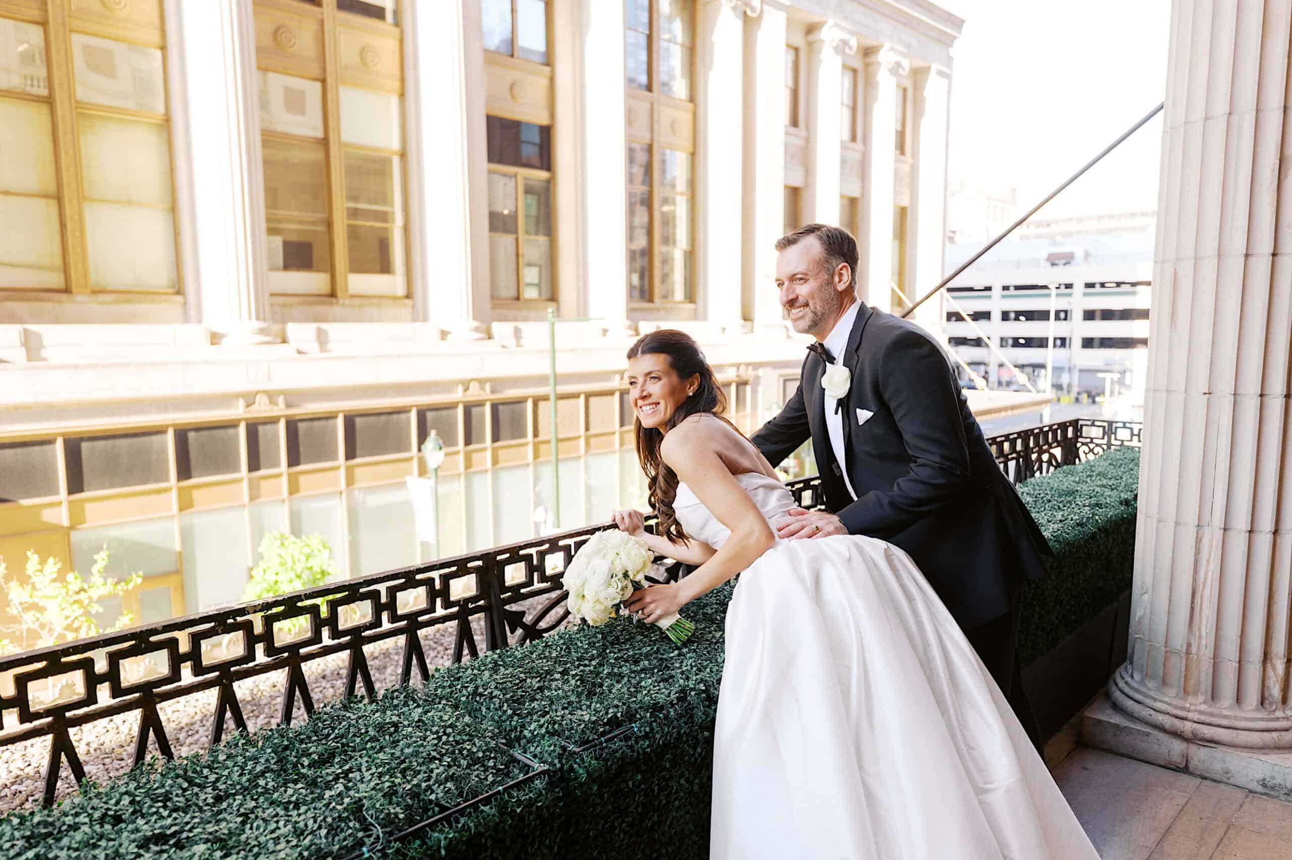 A bride and groom in formal attire smile and pose together on a balcony overlooking city buildings during the day, capturing the elegance of their classic wedding at Ballroom at the Ben.