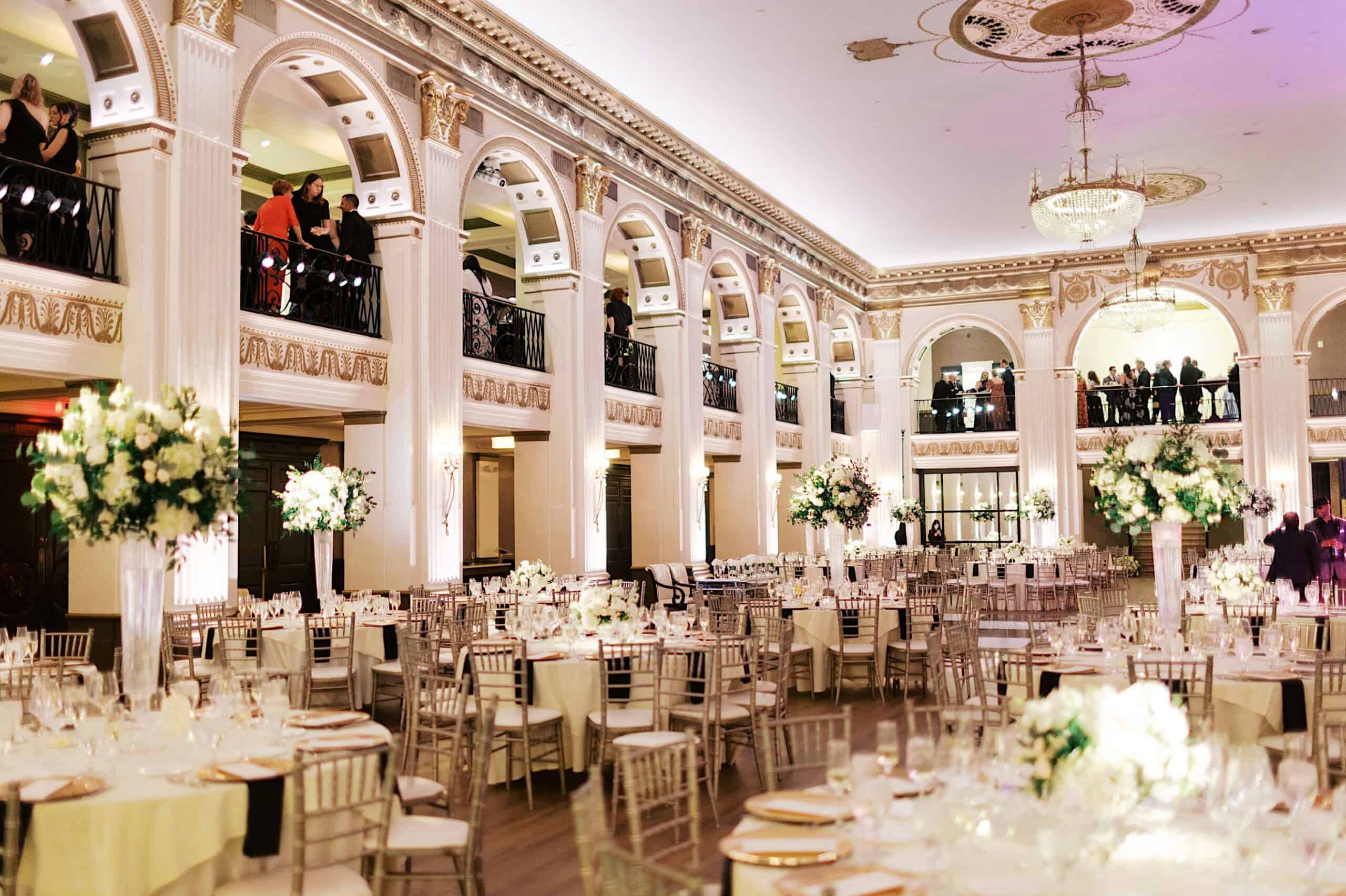 Elegant ballroom set for a formal event with round tables, gold chairs, tall floral centerpieces, and ornate architecture—perfect for a Classic Wedding at Ballroom at the Ben with sparkling chandeliers and charming balconies.