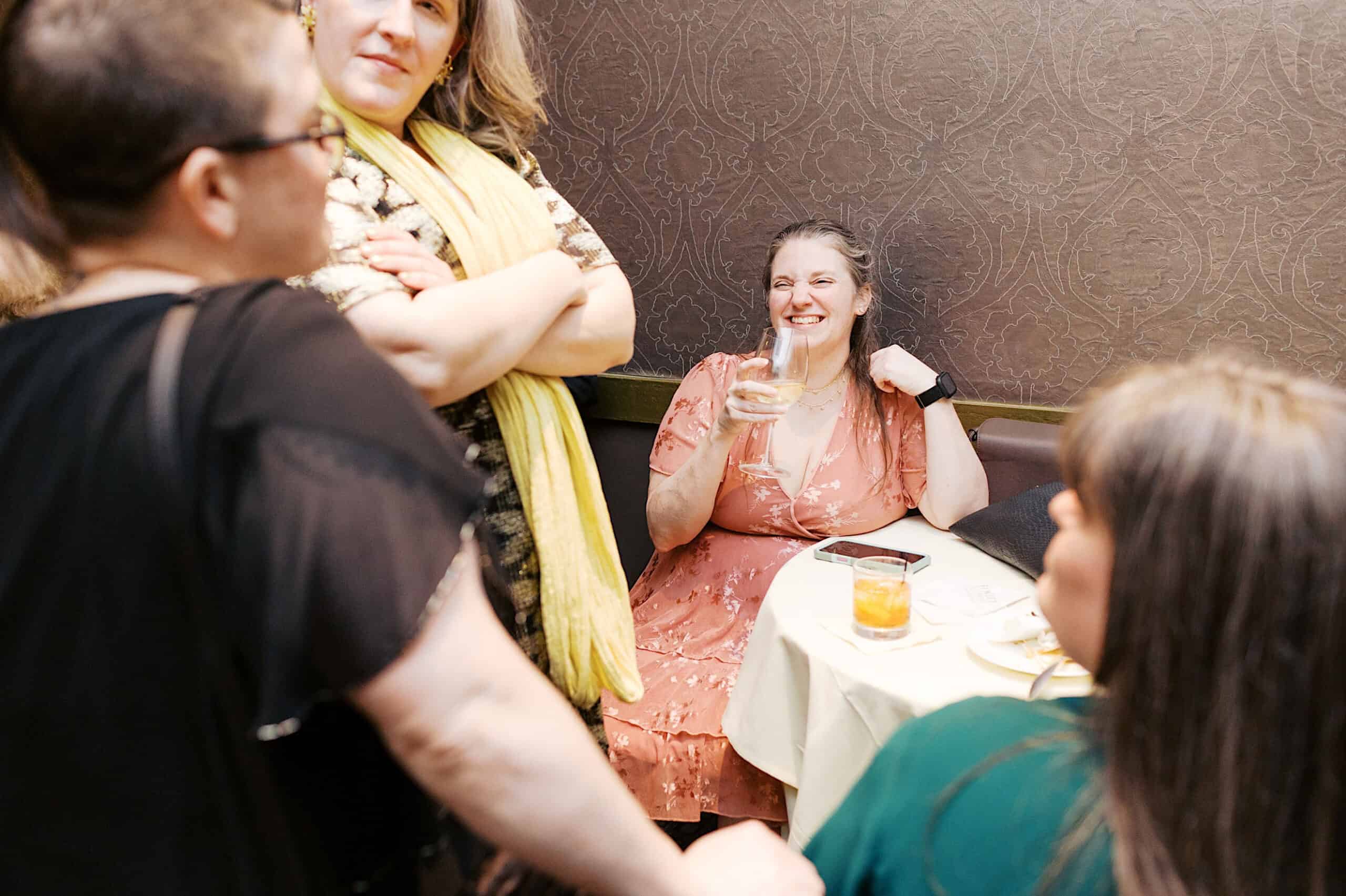 Four women gather around a small table in a dimly lit room at the Classic Wedding at Ballroom at the Ben; one woman is seated, smiling and holding a drink while others stand or sit nearby in lively conversation.