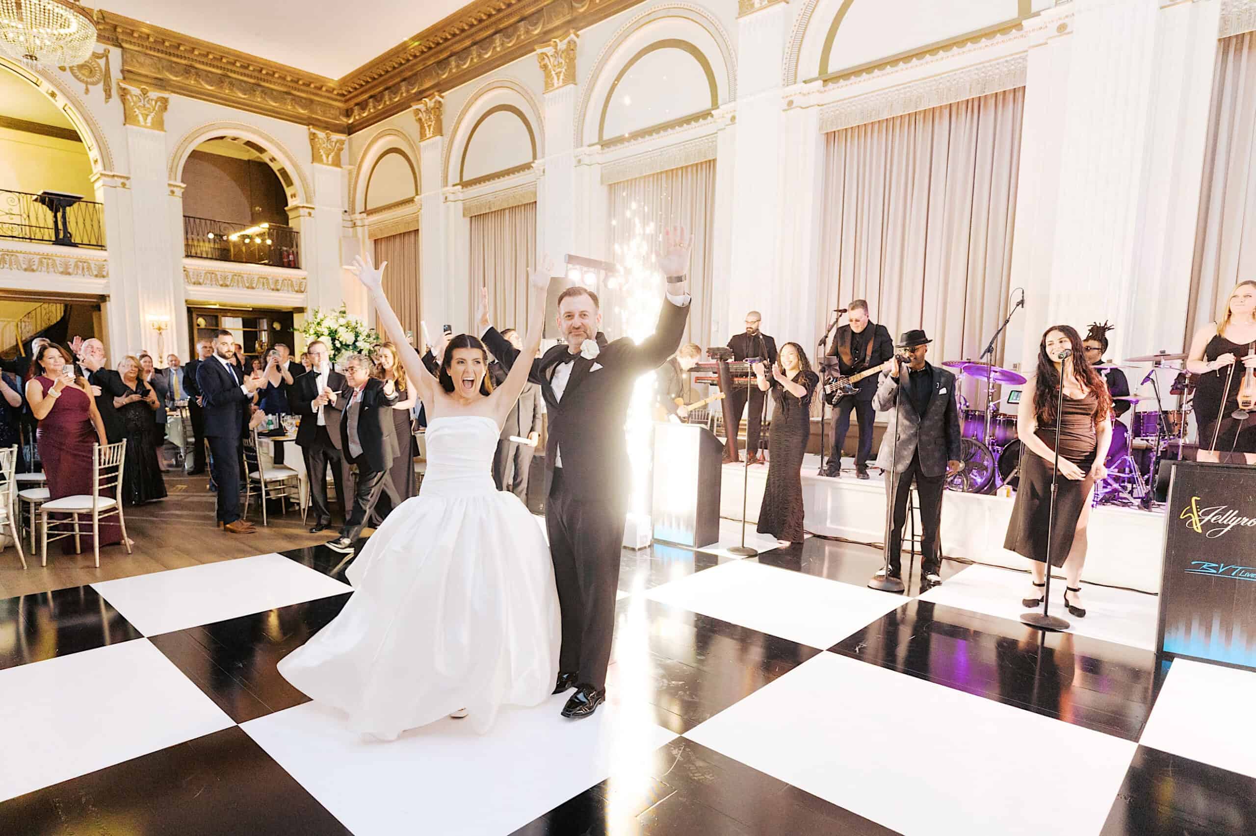 A bride and groom stand on a black and white checkered dance floor at their Classic Wedding at Ballroom at the Ben, raising their arms as guests applaud and a band performs in the background.