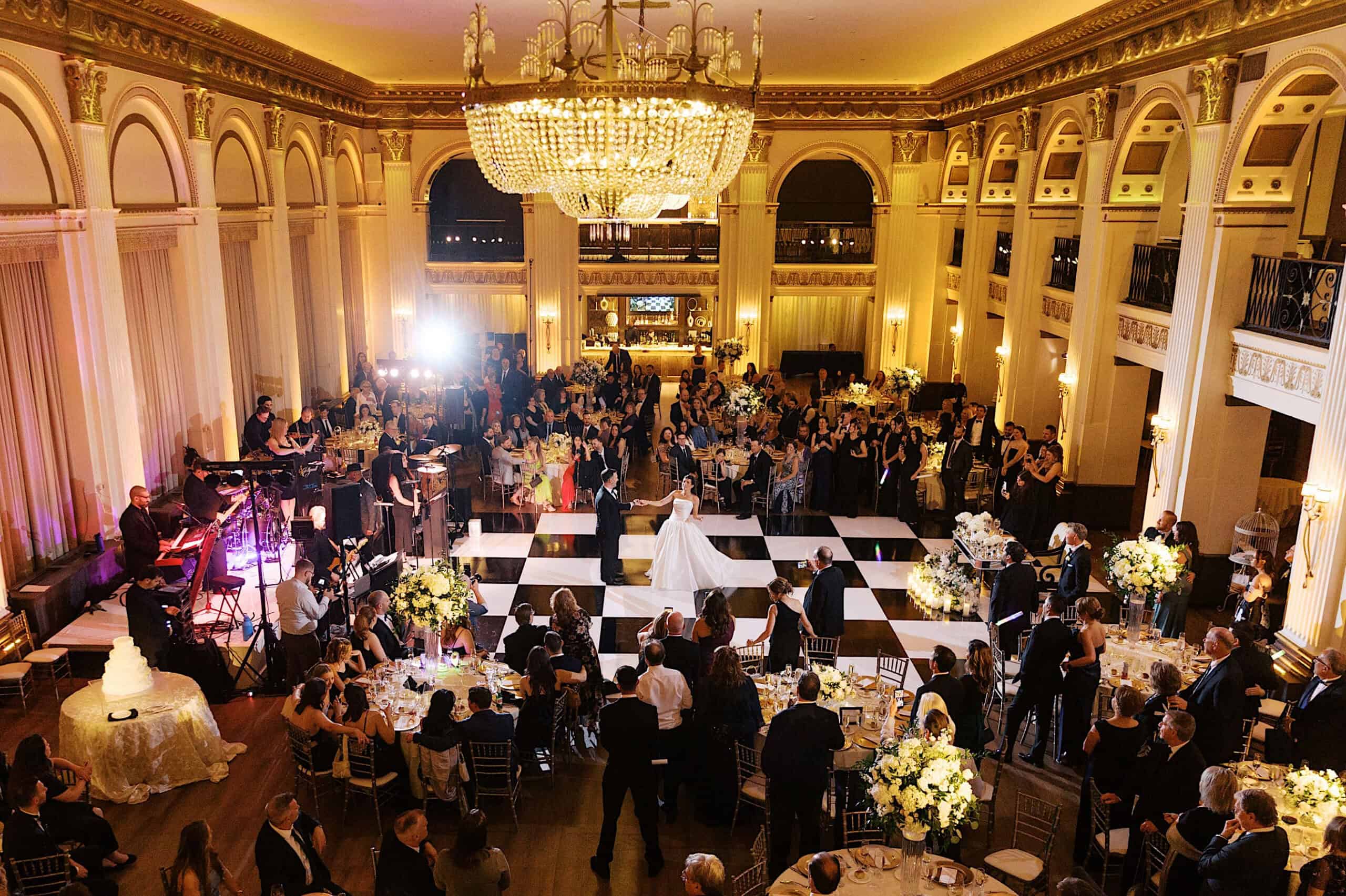A newlywed couple dances in the center of a large, elegant ballroom with checkered floor during their classic wedding at Ballroom at the Ben, surrounded by guests seated at decorated tables and a live band performing.