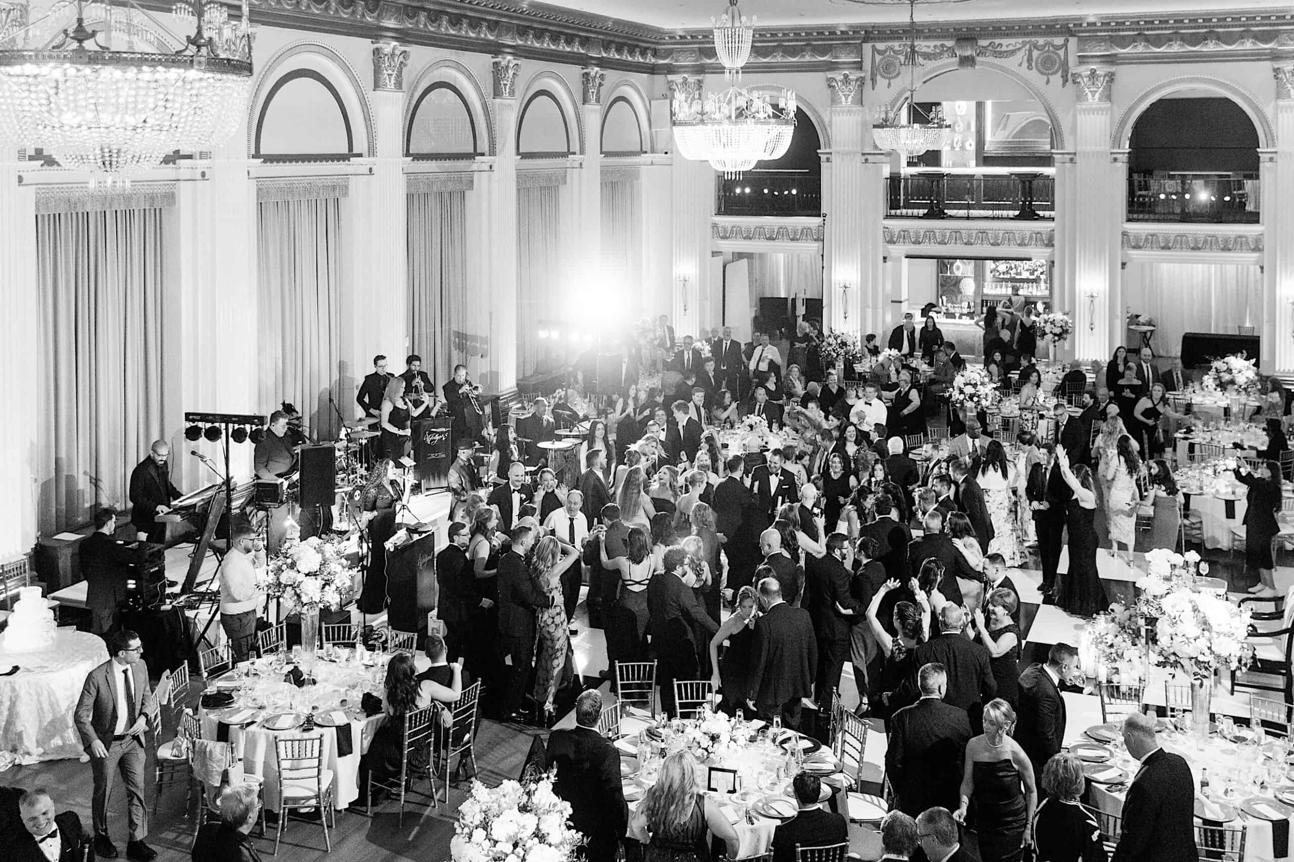 A large group of people in formal attire gathered for a Classic Wedding at Ballroom at the Ben, surrounded by decorated tables, chandeliers, and the sounds of a live band performing.