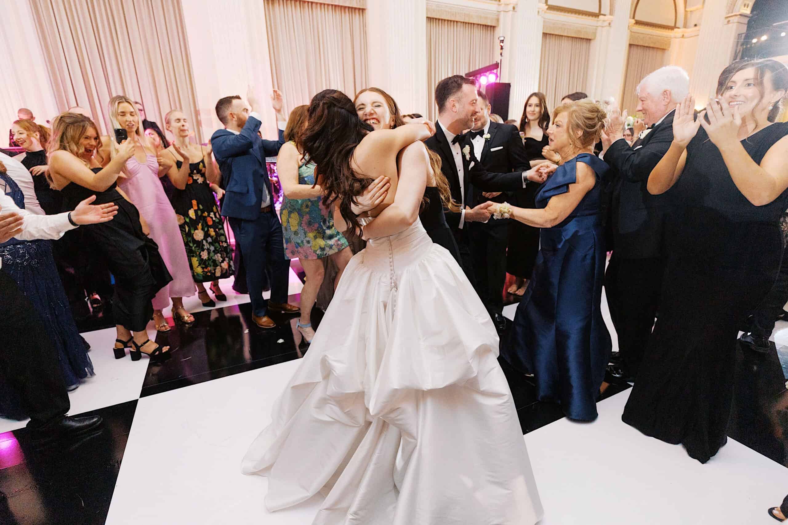 Two women in formal dresses embrace in the center of a dance floor during a classic wedding at Ballroom at the Ben, surrounded by well-dressed guests clapping and smiling at this elegant indoor event.
