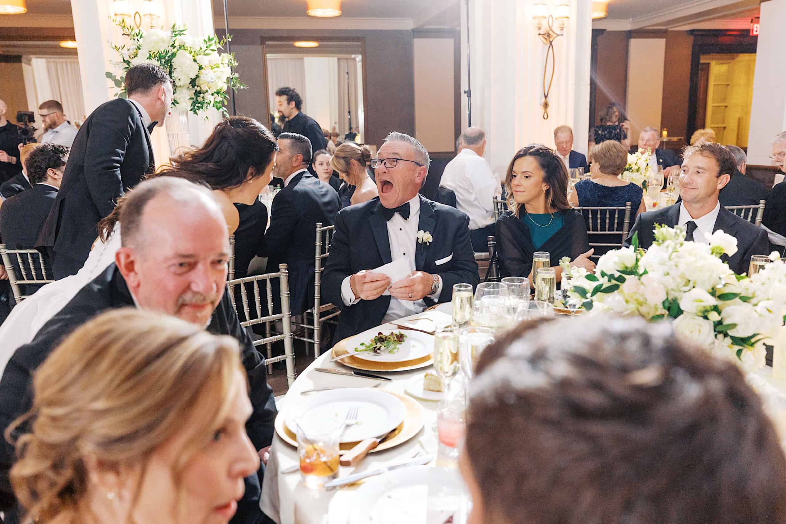 A group of people dressed formally sit around a decorated table at a Classic Wedding at Ballroom at the Ben, with one man in a tuxedo laughing animatedly.