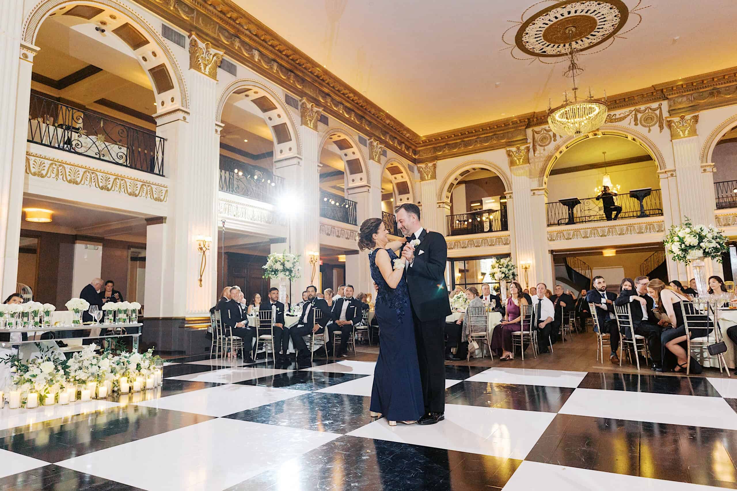 A couple dances in the center of a black-and-white checkered floor during a Classic Wedding at Ballroom at the Ben, surrounded by seated guests and elegant floral decorations.