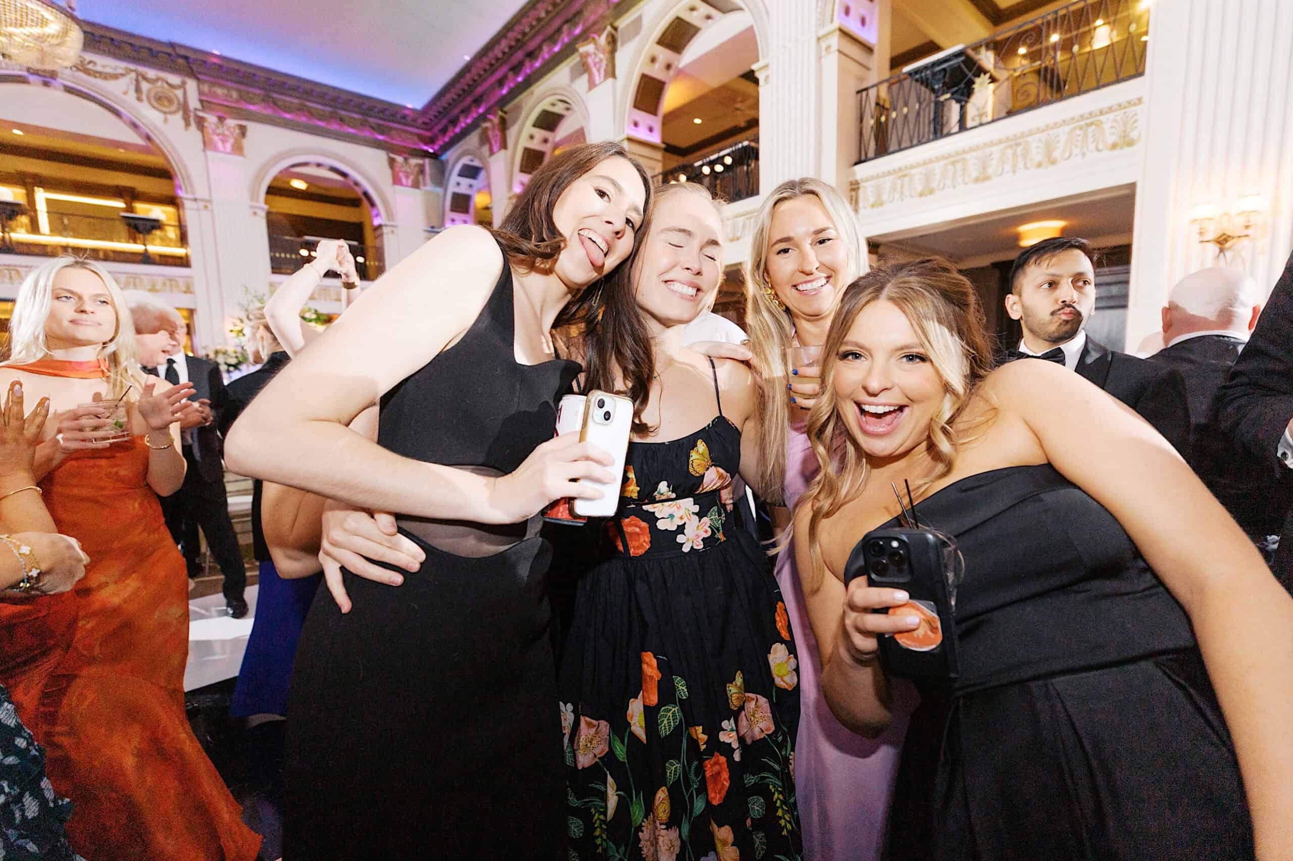 A group of women in formal attire smile and pose for a photo at a Classic Wedding at Ballroom at the Ben, holding drinks and phones, with other guests visible in the background.