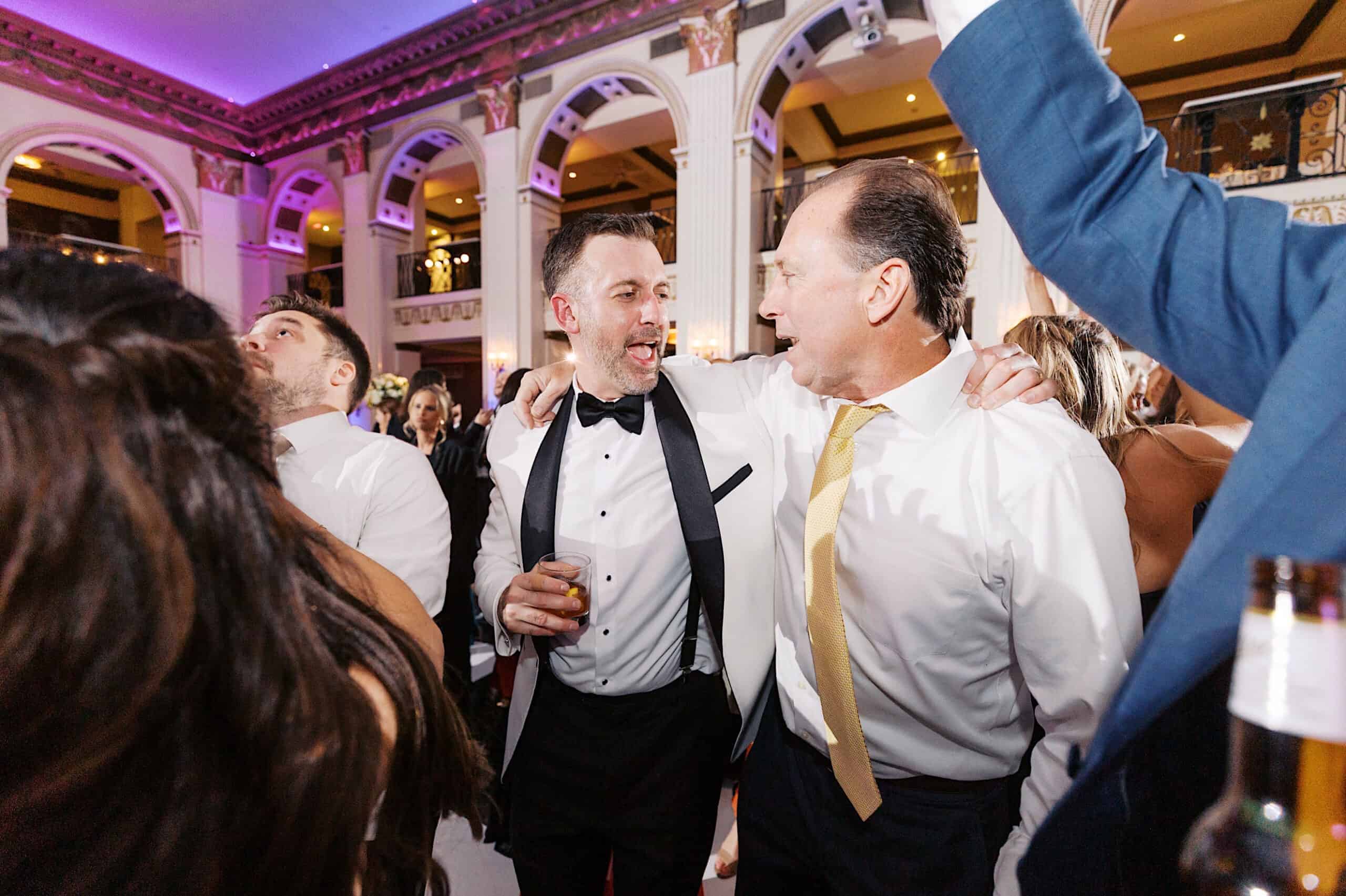 Two men in formal attire stand with arms around each other, smiling and talking at a lively Classic Wedding at Ballroom at the Ben, surrounded by guests and ornate architecture in the background.