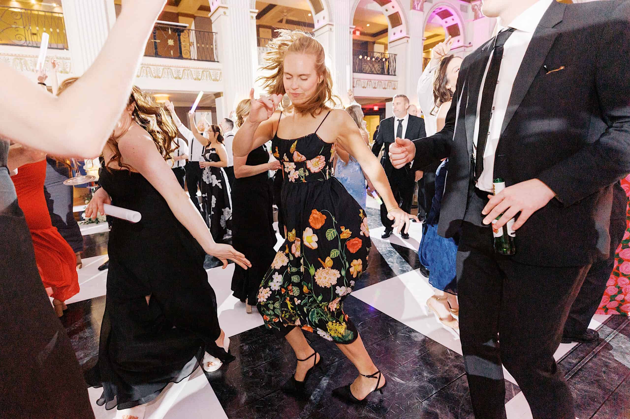 People in formal attire dance energetically on a black and white checkered floor at a Classic Wedding at Ballroom at the Ben, surrounded by ornate architecture and elegant balconies.