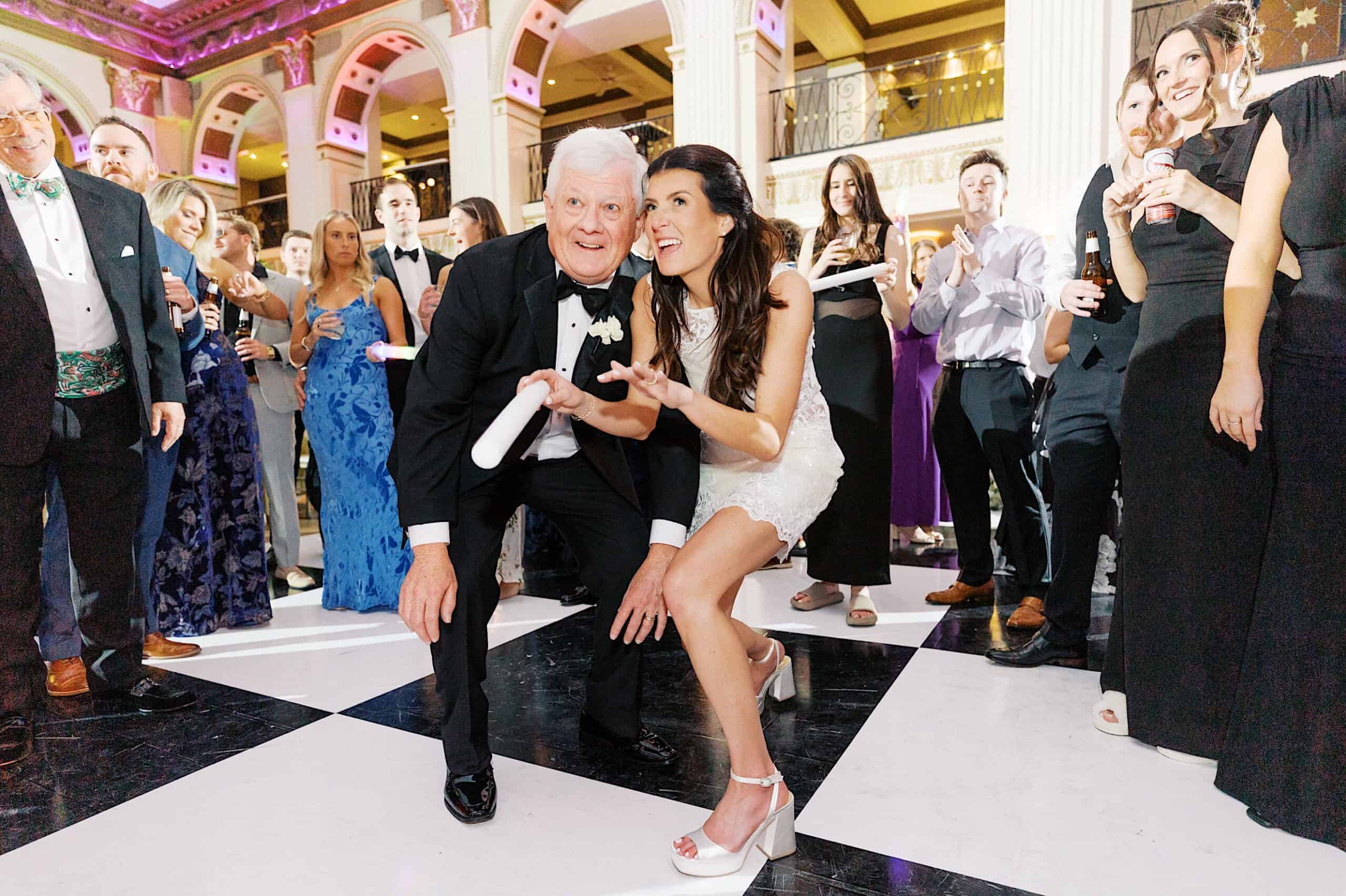 A bride in a white dress poses with an older man in a tuxedo on a black-and-white dance floor, surrounded by well-dressed guests at a Classic Wedding at Ballroom at the Ben.
