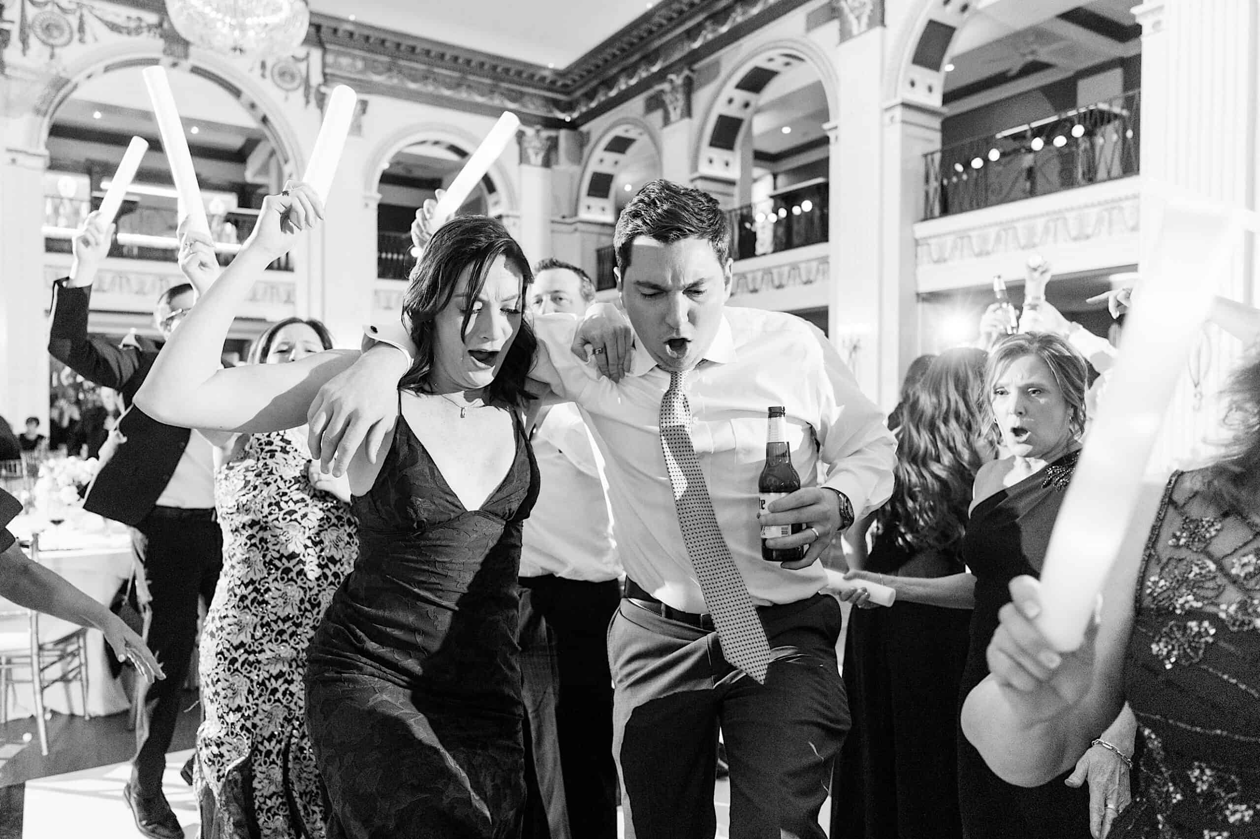 A group of people dance energetically at a Classic Wedding at Ballroom at the Ben, some waving light sticks. One man holds a beer bottle. The lively scene unfolds in an ornate and crowded ballroom.