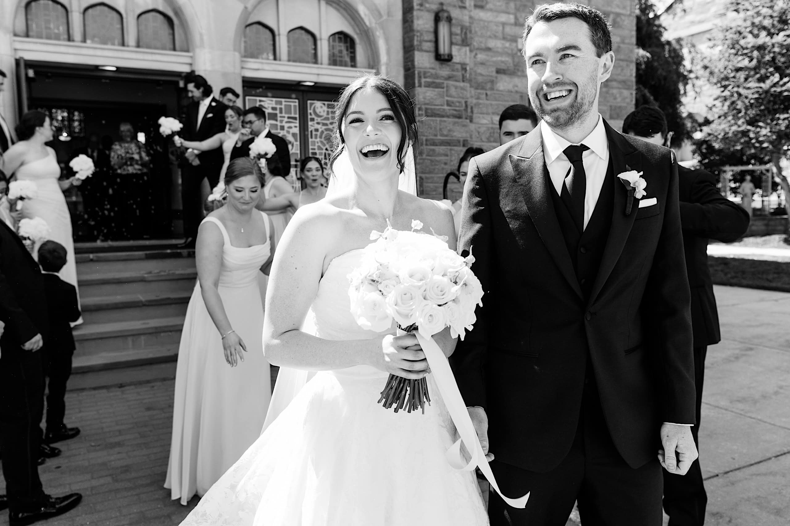 A bride and groom stand outside The Ivy at Ellis Preserve, smiling and holding hands, surrounded by wedding guests and bridesmaids with bouquets—a joyful spring wedding celebration.
