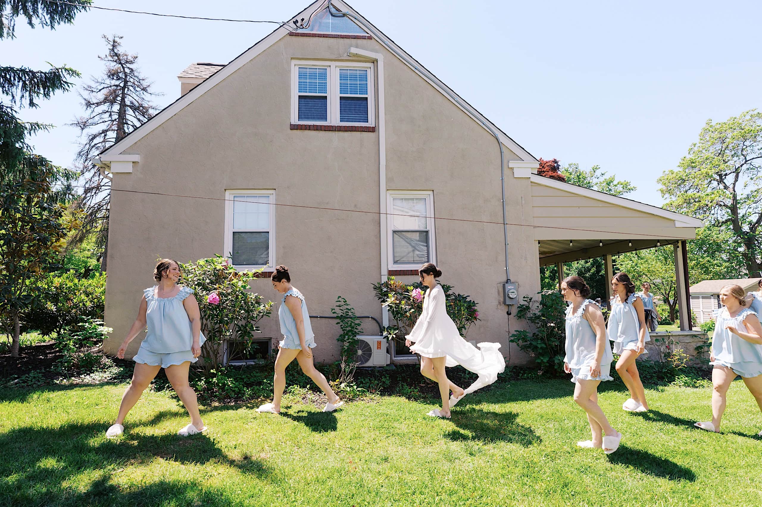 Six women wearing light blue outfits walk barefoot on grass in front of a beige house with a covered porch, capturing the joyful spirit of a The Ivy at Ellis Preserve spring wedding on a sunny day.