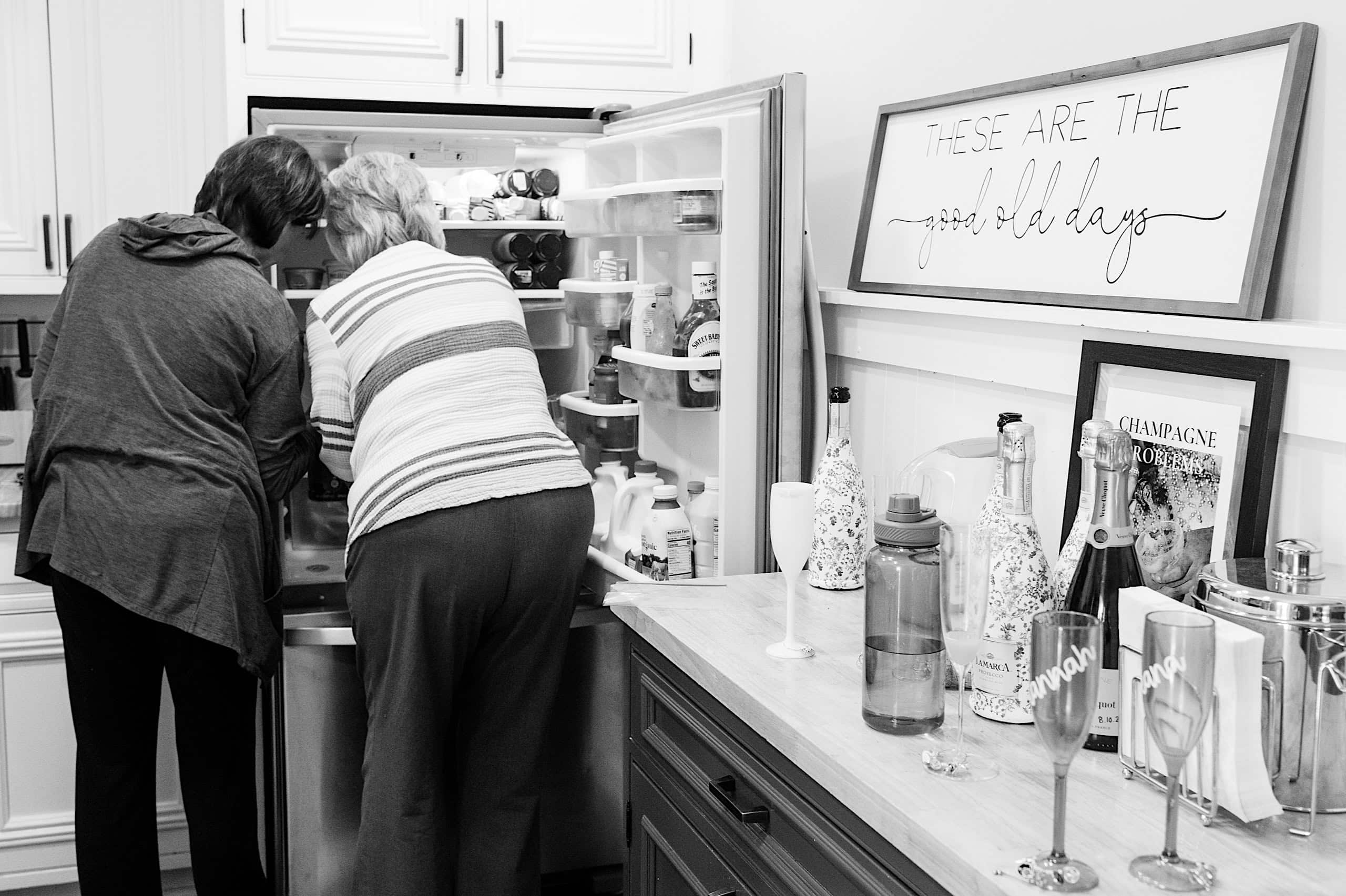 Two people stand side by side looking into an open refrigerator in a kitchen; a sign on the wall reads "These are the good old days," evoking nostalgia much like memories from The Ivy at Ellis Preserve spring wedding.