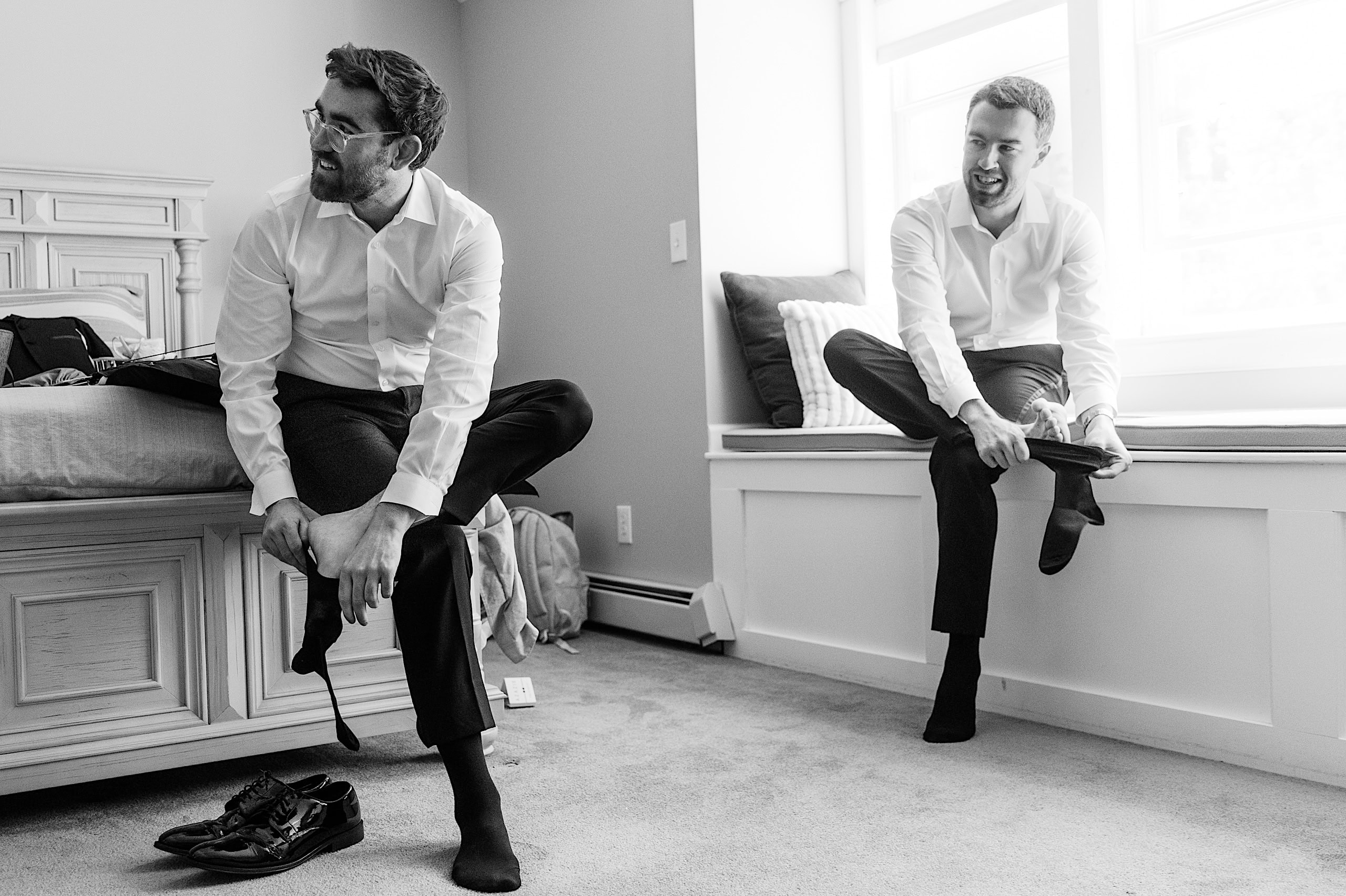Two men in dress shirts and pants sit putting on socks in a bright bedroom at The Ivy at Ellis Preserve, one on the bed and one on a window seat, with wedding shoes and clothing nearby, preparing for a spring wedding.