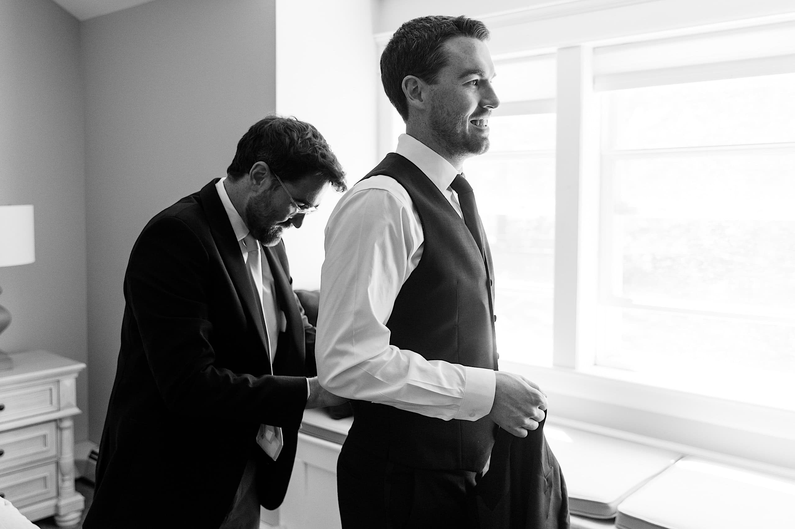 Two men in formal attire prepare for an event in a bright room at The Ivy at Ellis Preserve; one helps the other with his suit jacket before a spring wedding.