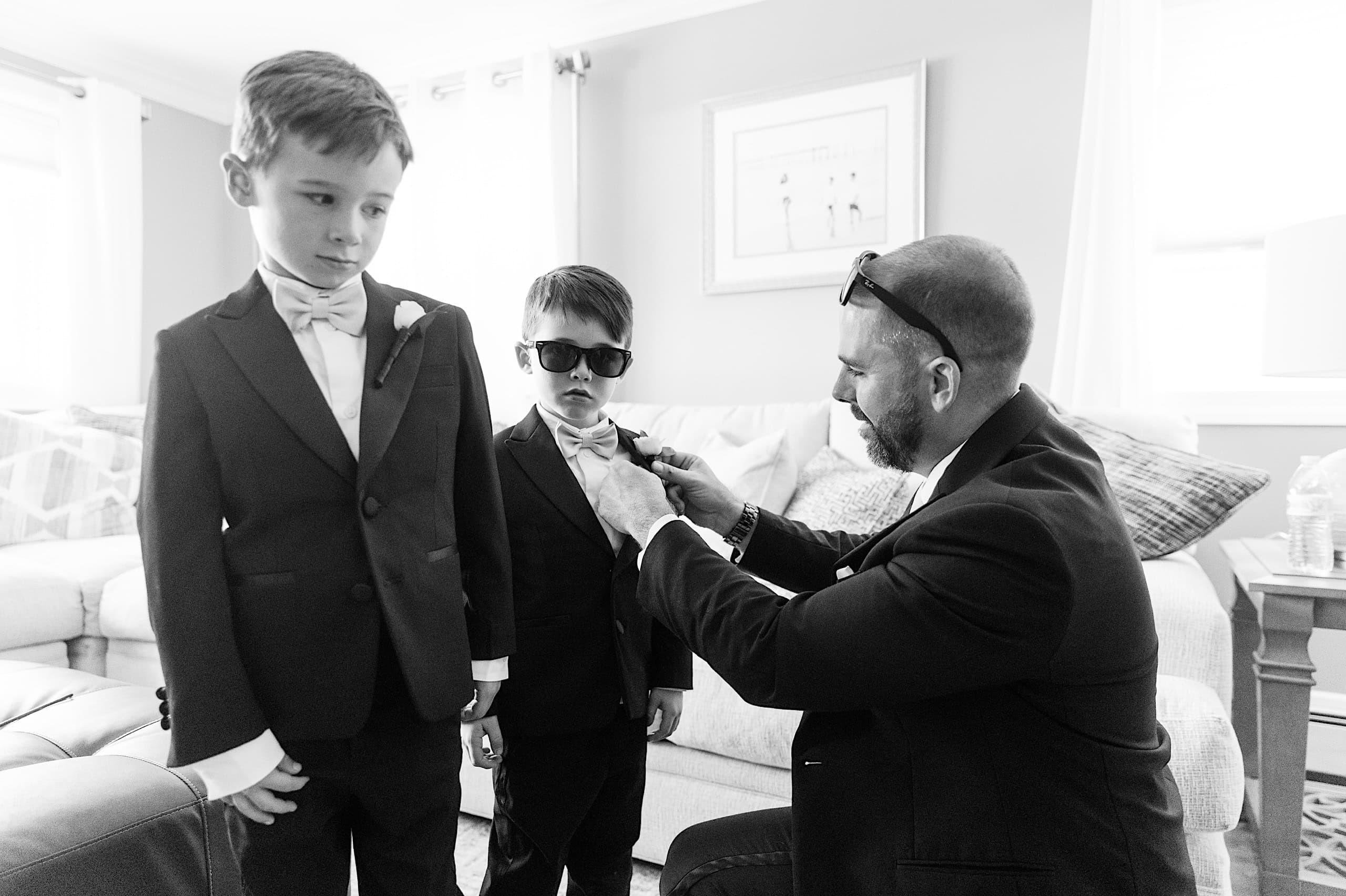 An adult helps adjust the bowtie of a young boy in a suit and sunglasses, while another suited boy stands beside them in a living room, capturing sweet moments before The Ivy at Ellis Preserve spring wedding.