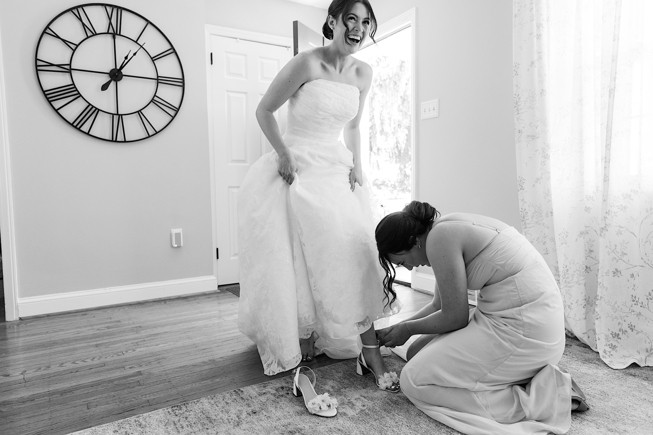 A bride smiles while standing at her The Ivy at Ellis Preserve spring wedding as another woman kneels to help fasten her shoe in a bright room with a large wall clock.