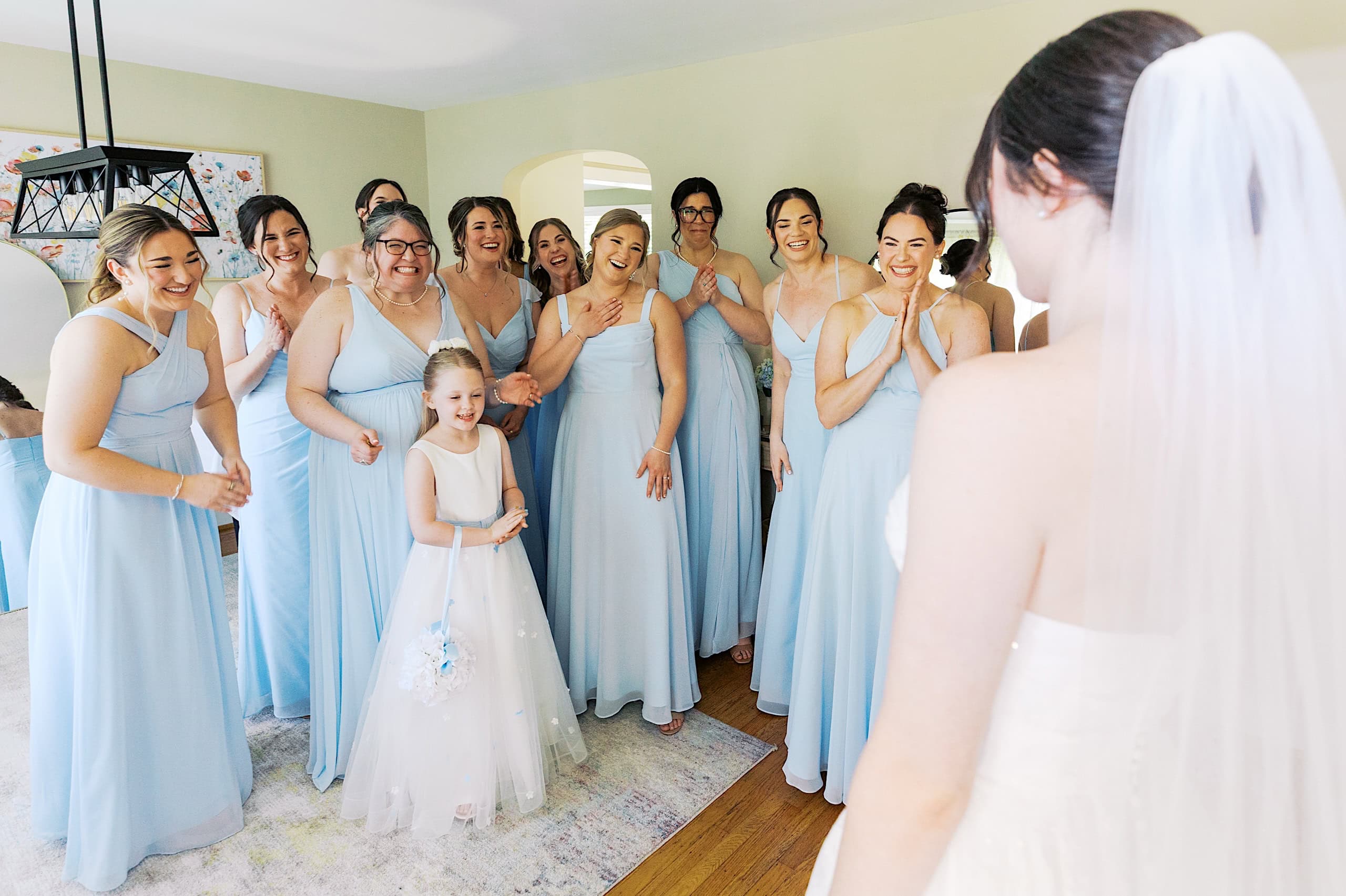 A bride in a white gown faces joyful bridesmaids and a flower girl in light blue dresses, all beaming inside a bright room at The Ivy at Ellis Preserve during a beautiful spring wedding.