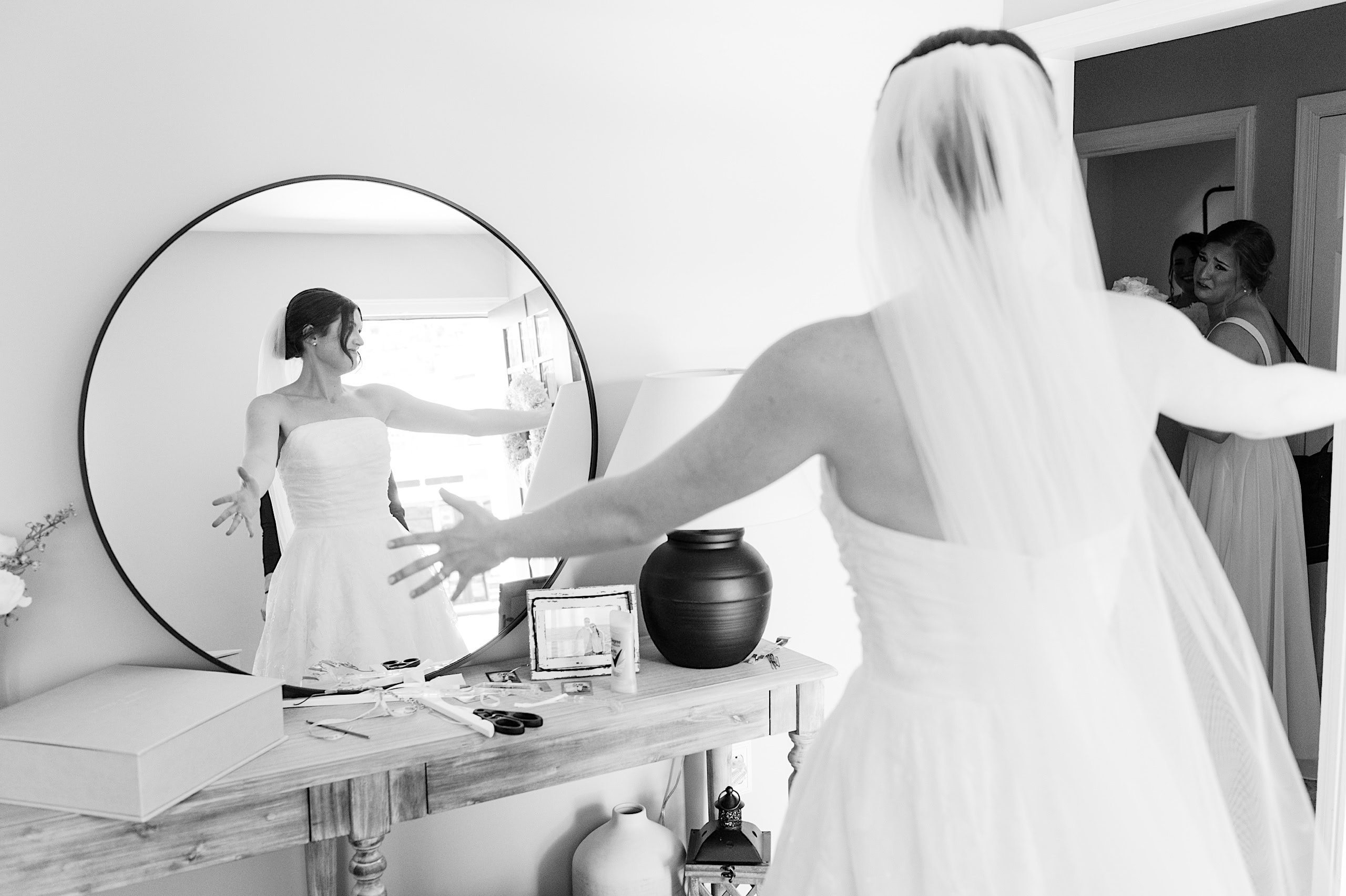 A bride in a wedding dress stands with arms outstretched in front of a mirror at The Ivy at Ellis Preserve spring wedding, her reflection visible, while another woman looks on from a doorway.