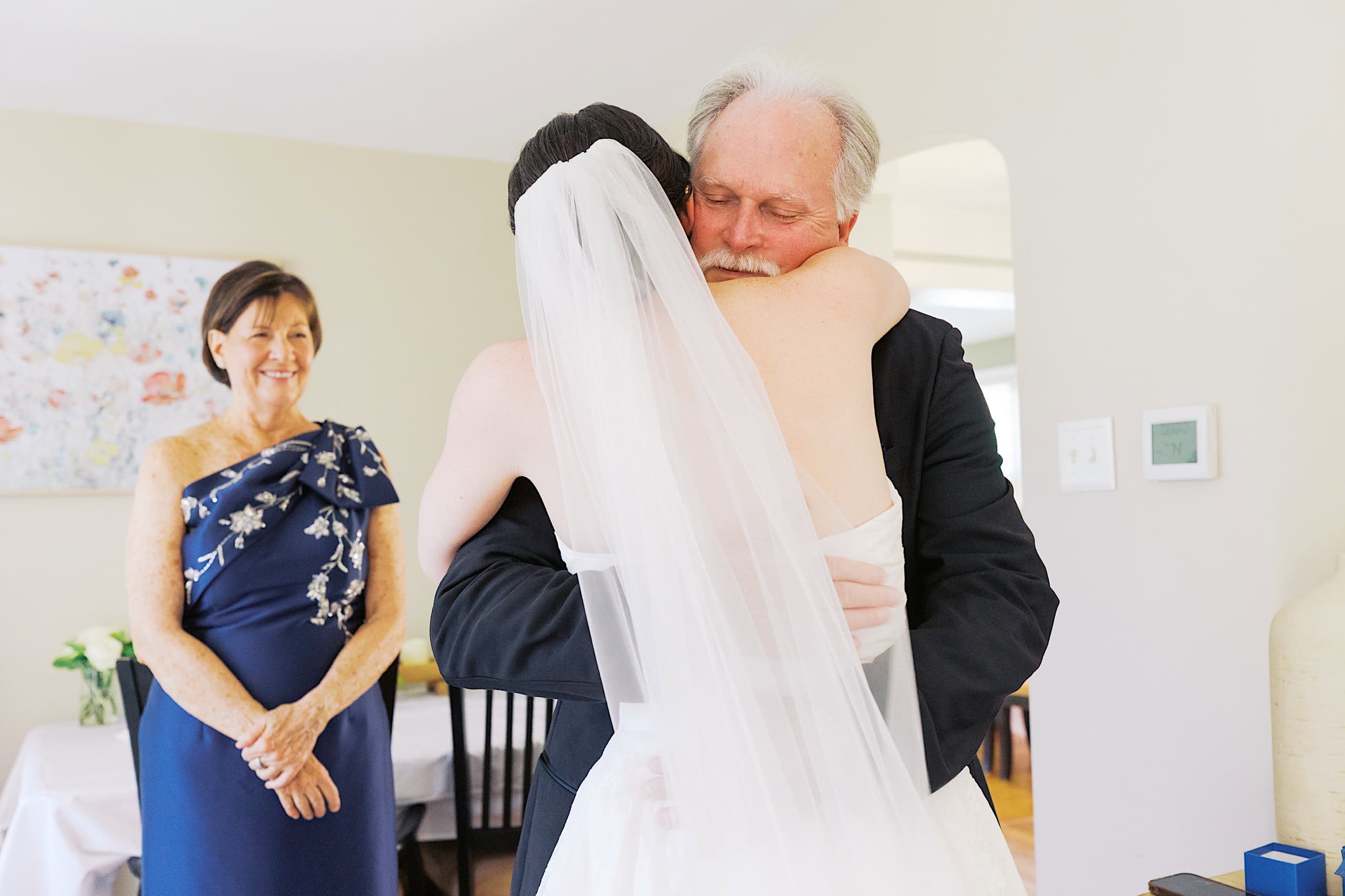 A bride in a white dress and veil hugs an older man in a suit, while an older woman in a blue dress smiles nearby at The Ivy at Ellis Preserve spring wedding.