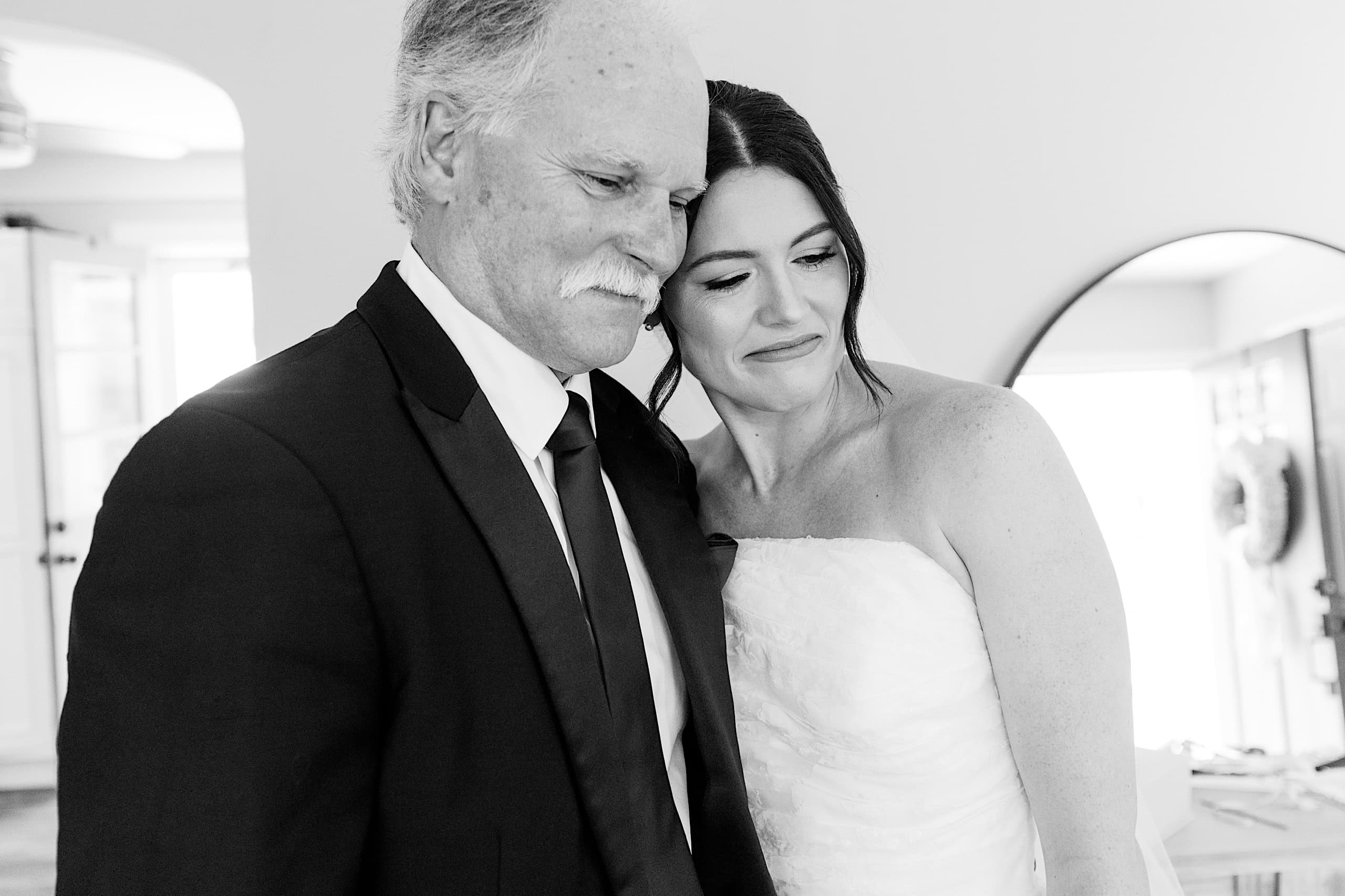 A woman in a wedding dress stands closely beside an older man in a suit; both appear emotional in the bright indoor setting of The Ivy at Ellis Preserve spring wedding.