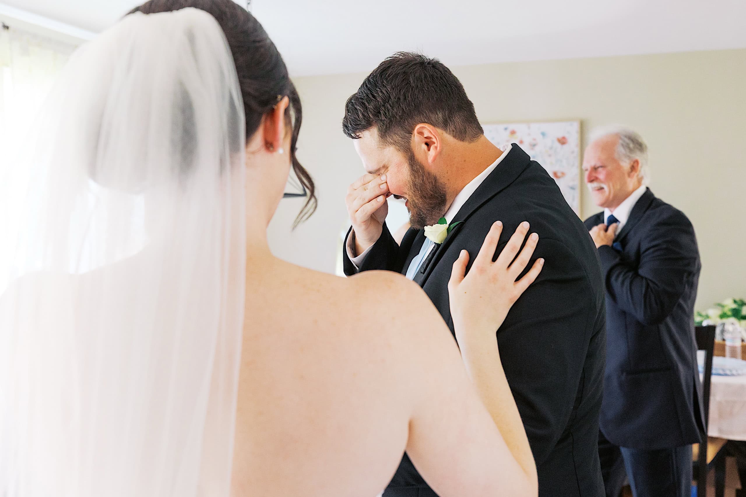 At a beautiful The Ivy at Ellis Preserve spring wedding, a bride touches the shoulder of a groom wiping his eyes, while an older man in a suit smiles in the background.