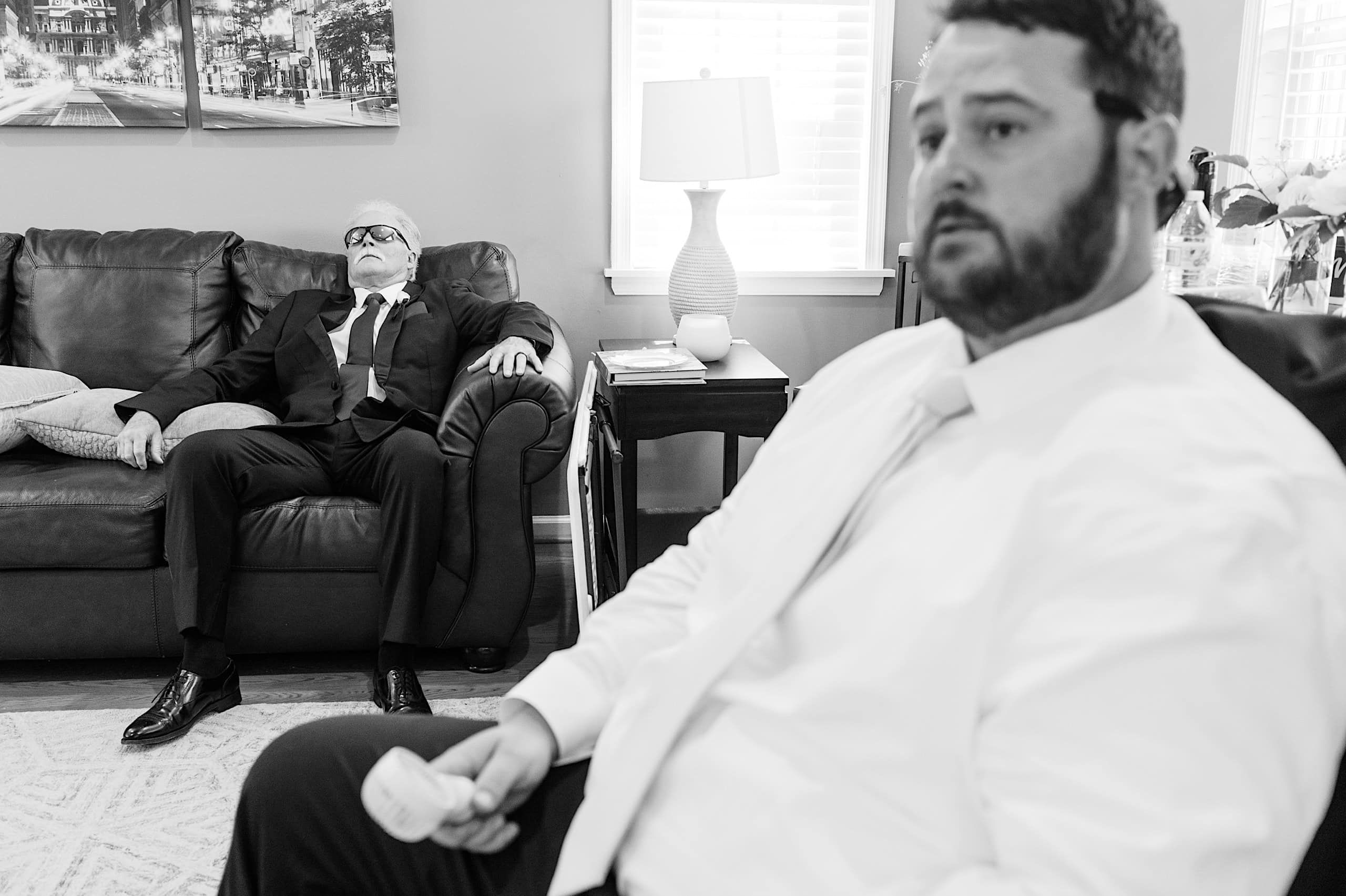 Two men in formal attire are in a living room; one is sitting on a couch with eyes closed, while the other sits in the foreground holding an object, preparing for The Ivy at Ellis Preserve spring wedding.