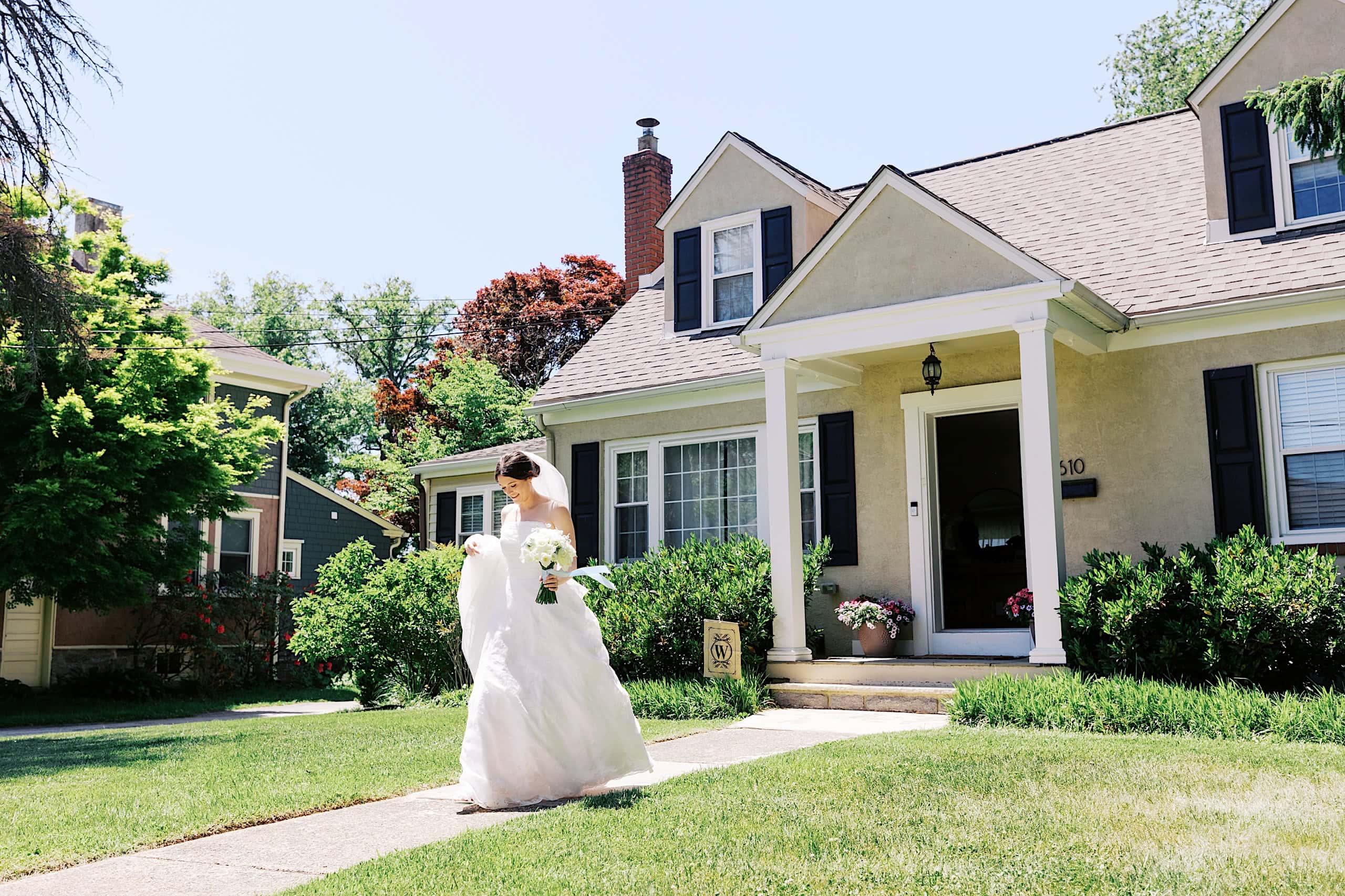 A bride in a white wedding dress holding a bouquet walks on a sidewalk in front of a beige suburban house on a sunny day, capturing the joy and elegance of The Ivy at Ellis Preserve spring wedding.