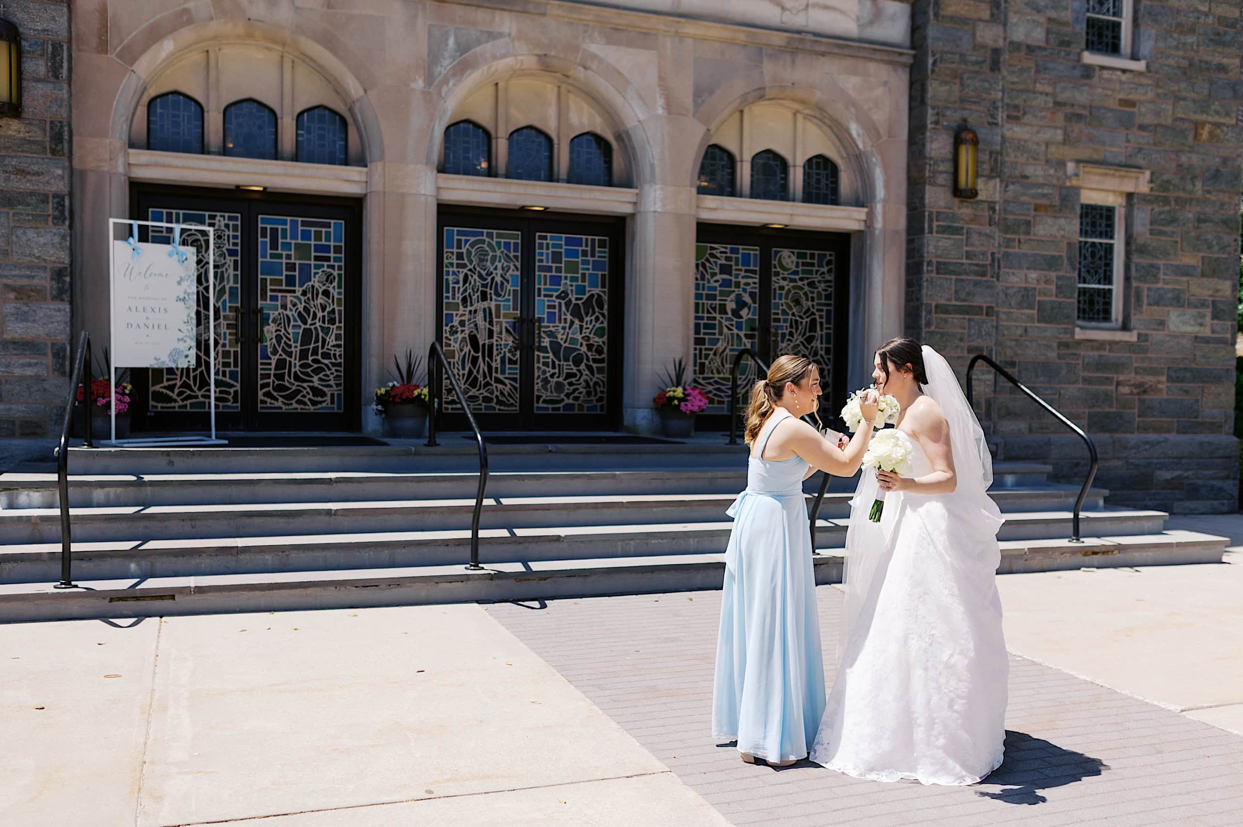 A woman in a light blue dress adjusts the veil of a bride in a white gown, standing outside the stained glass doors of The Ivy at Ellis Preserve during a beautiful spring wedding.