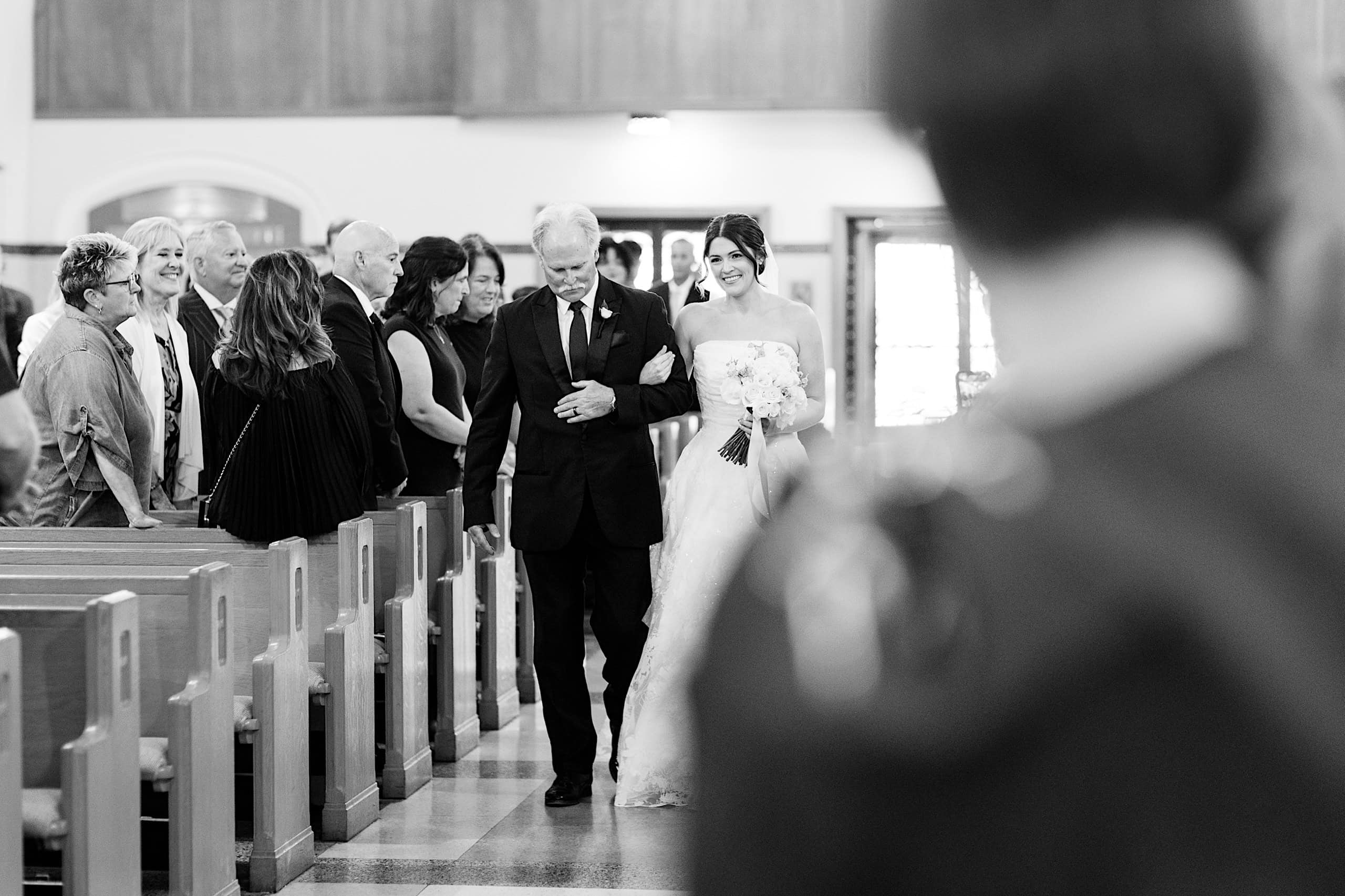 A bride in a wedding dress walks down the aisle with her father at The Ivy at Ellis Preserve spring wedding, while guests seated in pews watch in a beautiful church setting.