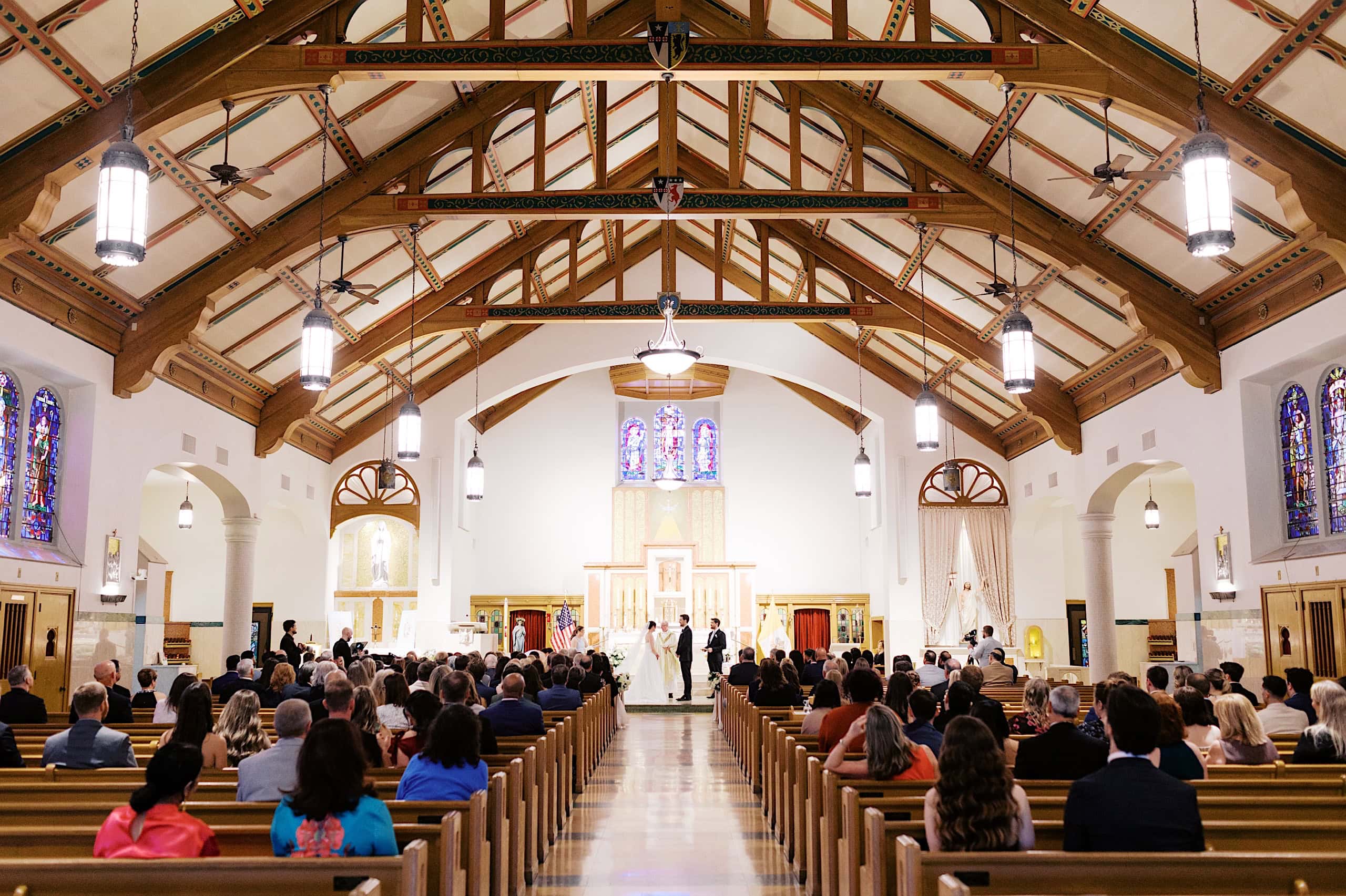 A wedding ceremony takes place at the front of a church, with guests seated in pews facing the altar and stained glass windows along the sides, evoking the elegance of The Ivy at Ellis Preserve spring wedding.