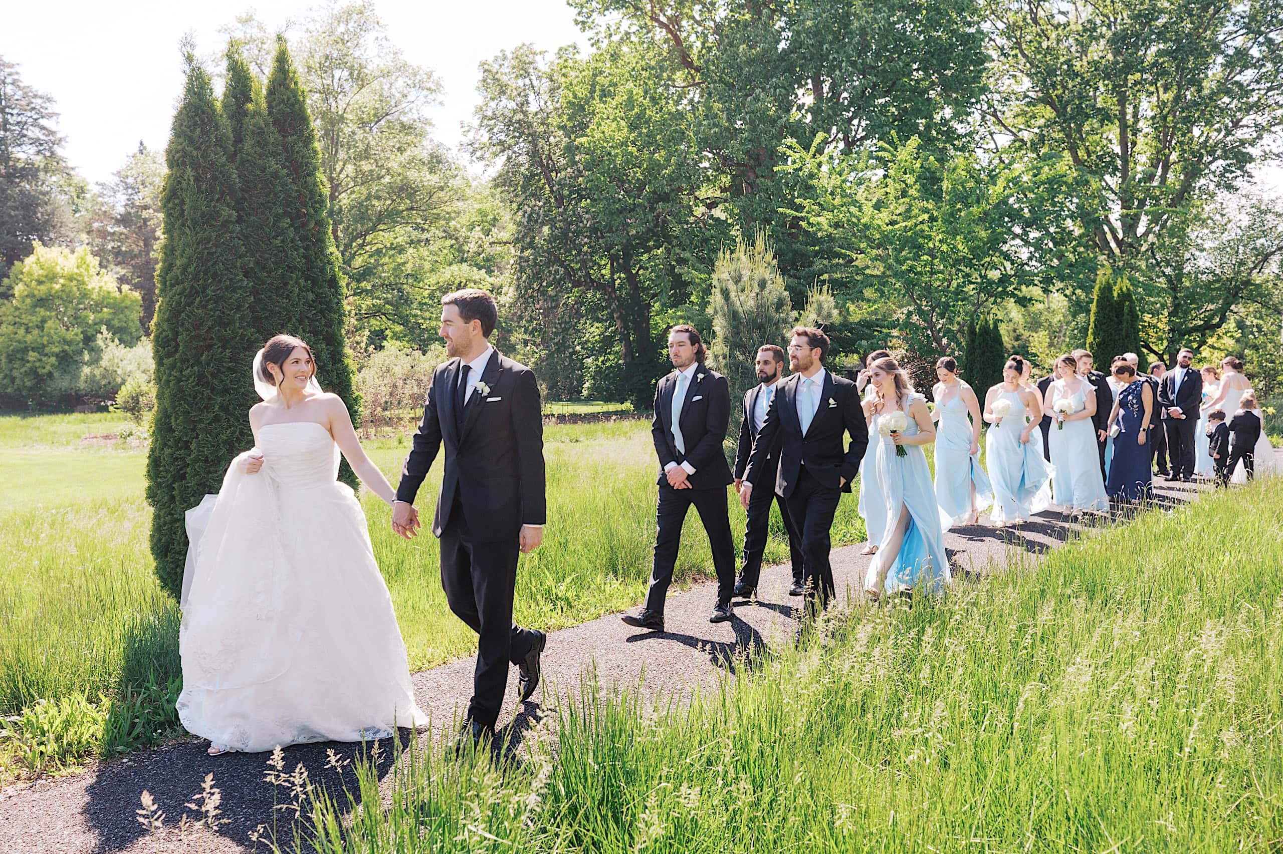 A bride and groom walk hand in hand along a path at The Ivy at Ellis Preserve spring wedding, followed by their wedding party dressed in formal attire amidst greenery and trees.