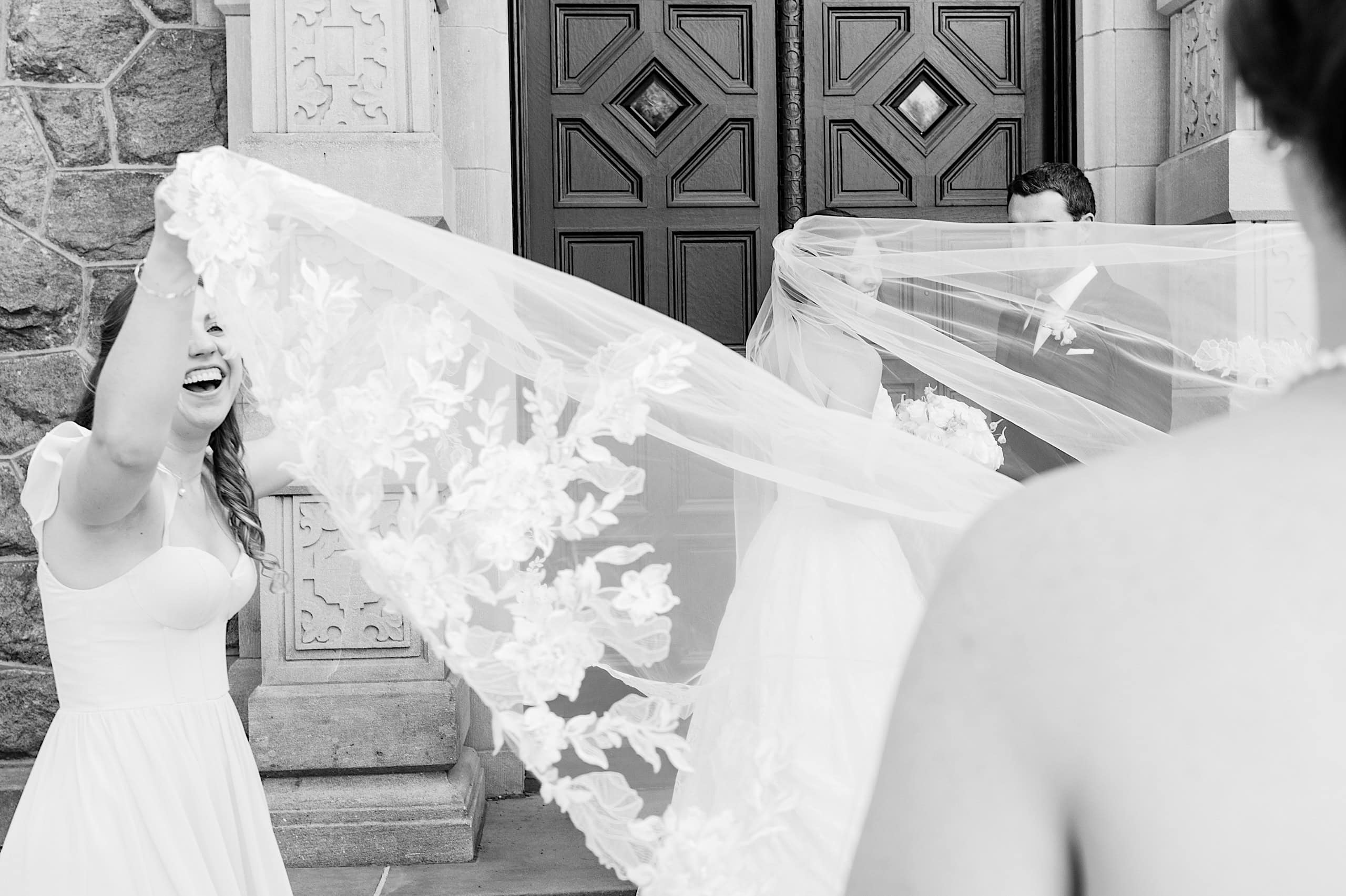 At The Ivy at Ellis Preserve spring wedding, a woman holds up a bride's long lace veil outside the building, while another person and a man in formal attire stand nearby.