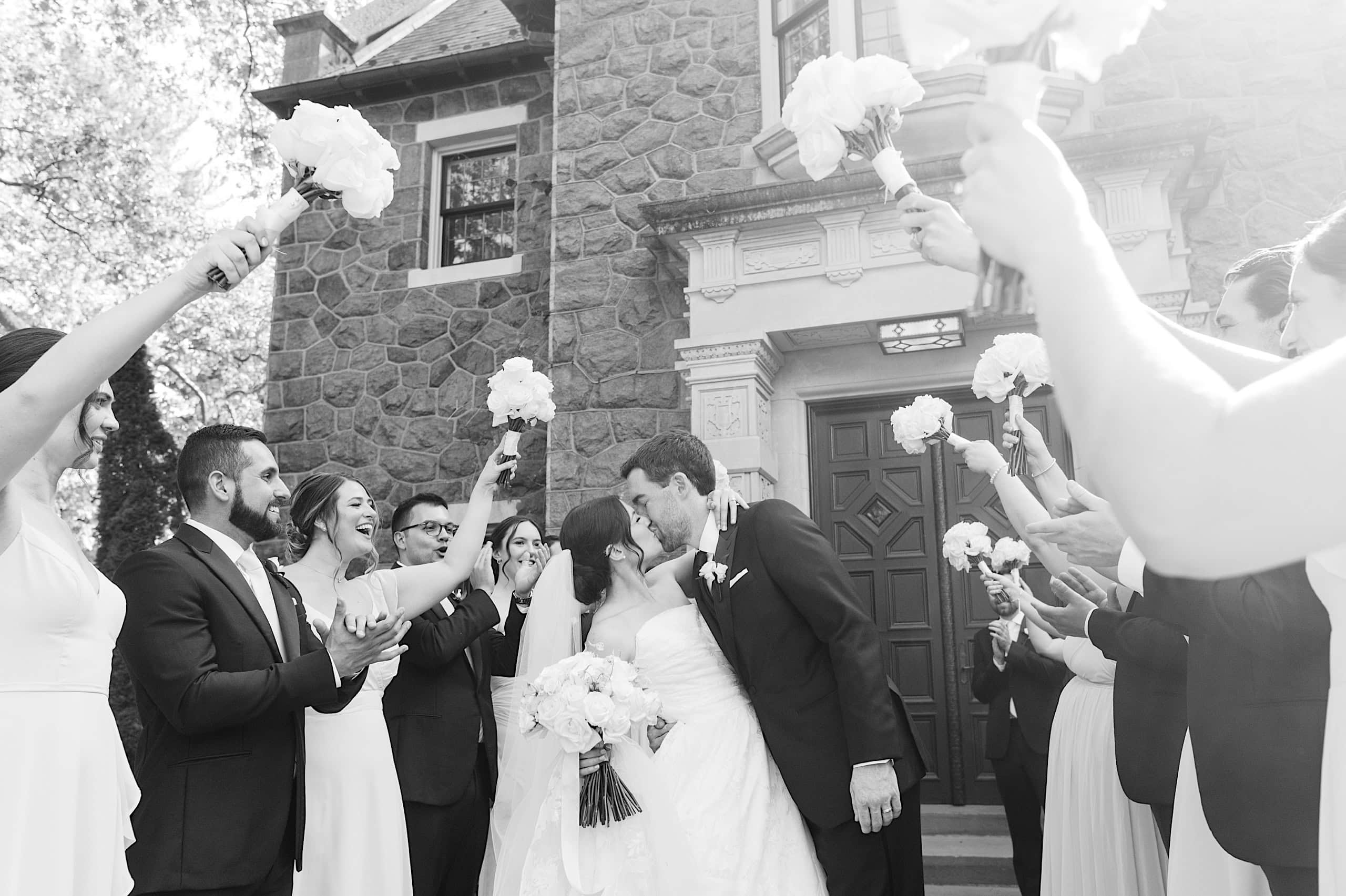 A bride and groom kiss outside a stone building at The Ivy at Ellis Preserve spring wedding, while guests stand on either side, holding bouquets and clapping.