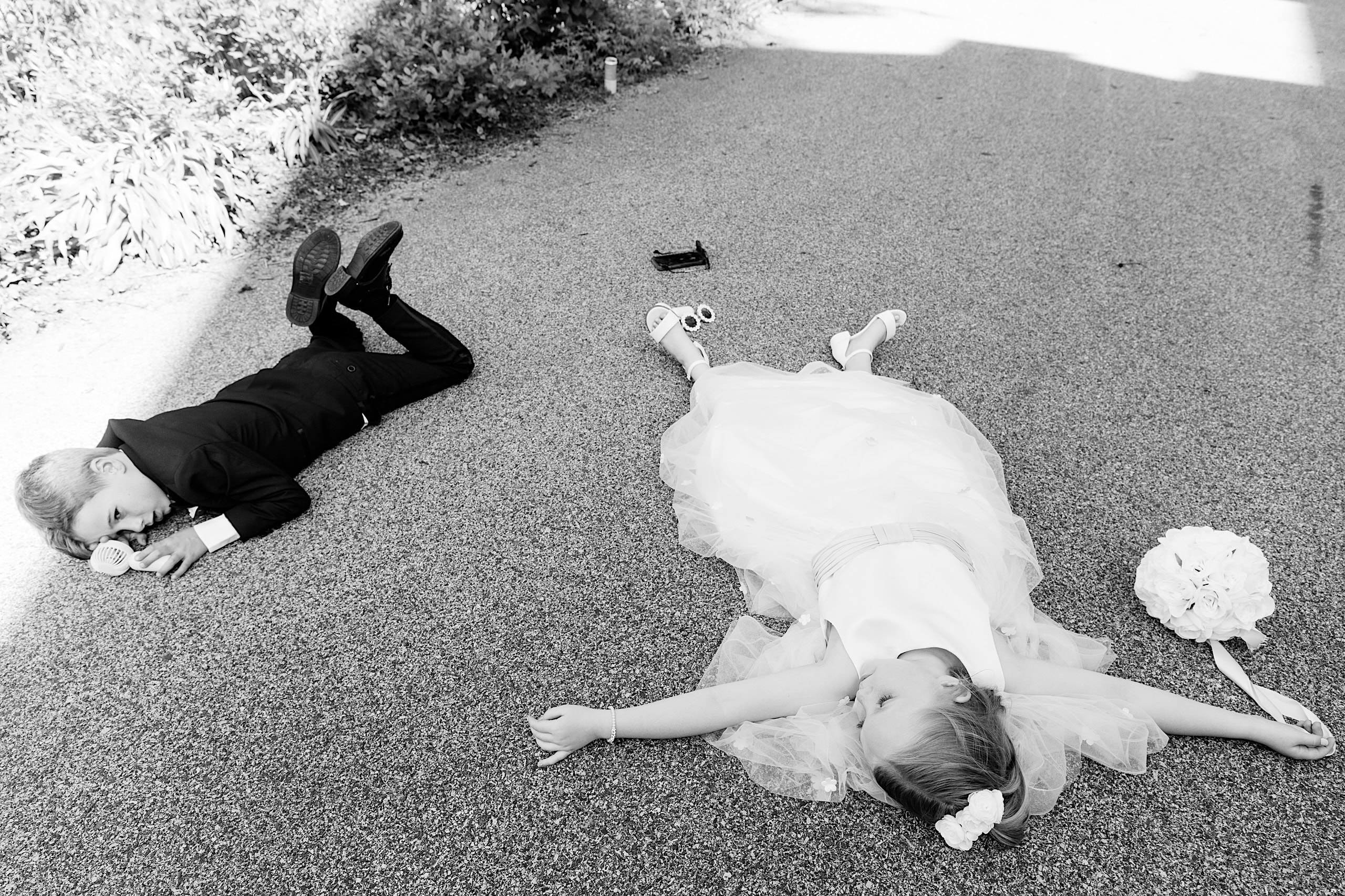 A boy in a suit and a girl in a dress lie on the ground, arms and legs outstretched as if exhausted after The Ivy at Ellis Preserve spring wedding; a bouquet and toy car rest nearby.