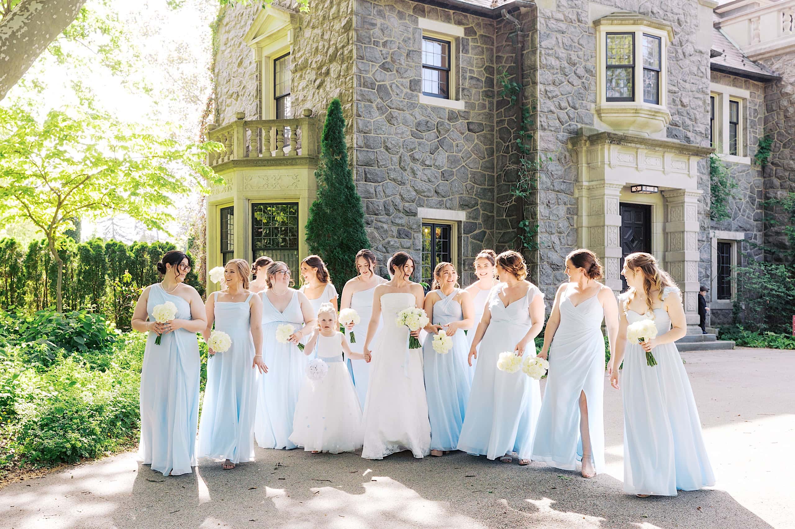 A bride and her bridesmaids in light blue dresses stand outside The Ivy at Ellis Preserve, holding white bouquets and smiling in the sunlight during a beautiful spring wedding.