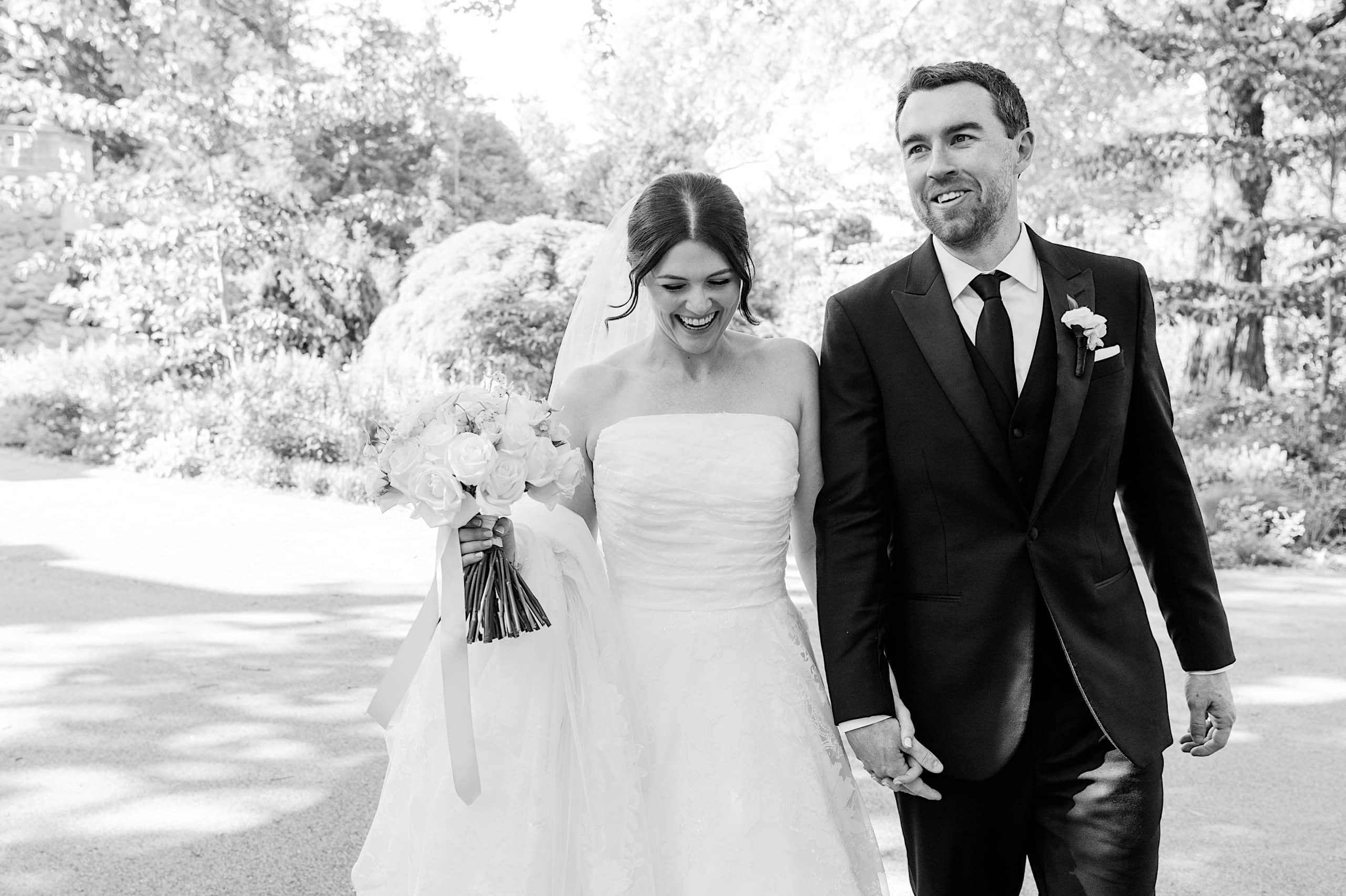 A bride and groom walk outdoors, holding hands and smiling during their The Ivy at Ellis Preserve spring wedding. The bride wears a strapless gown with a bouquet; the groom dons a dark suit with a boutonnière.