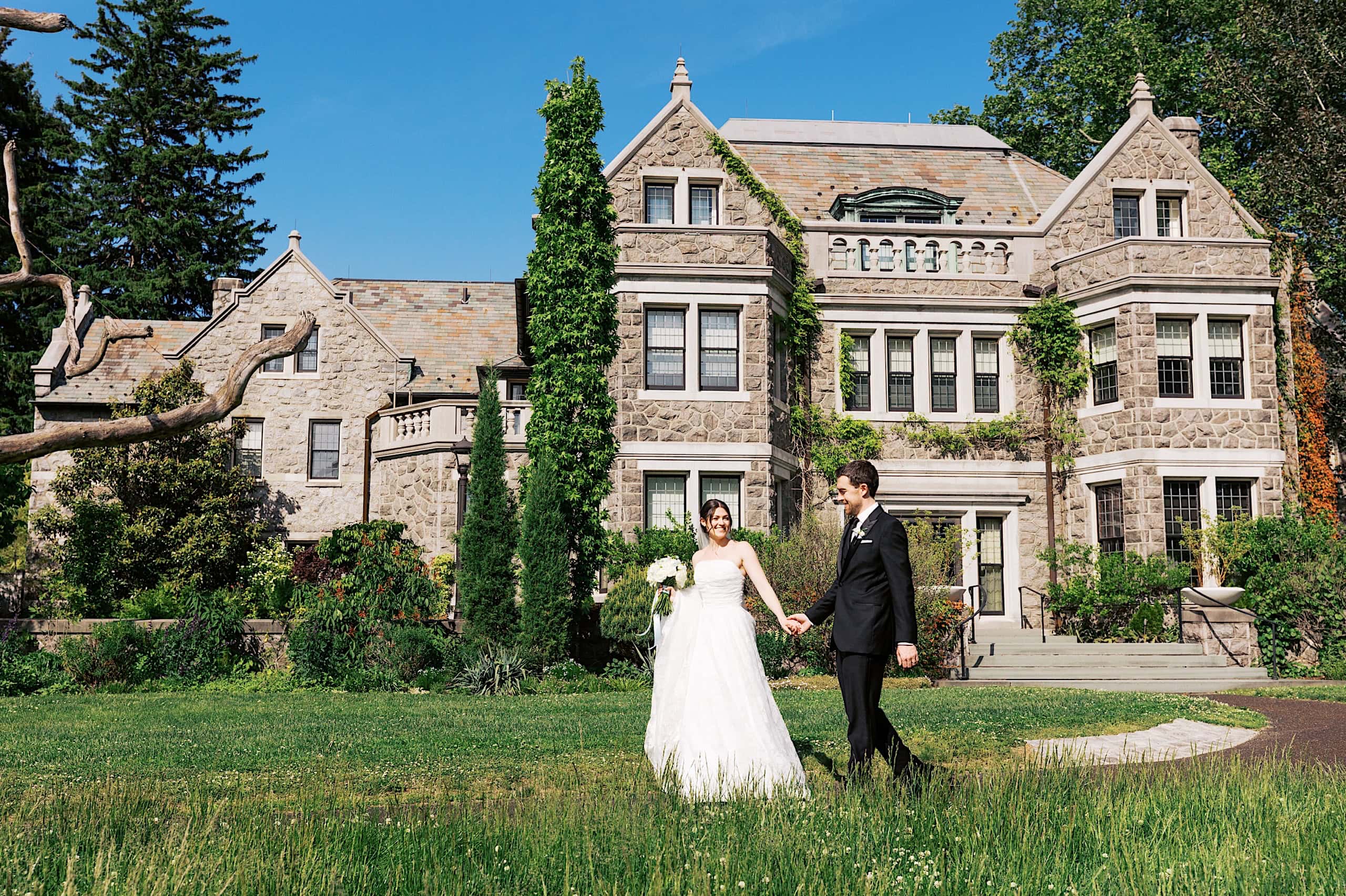 A bride and groom walk hand in hand on the lawn in front of a large stone mansion during their The Ivy at Ellis Preserve spring wedding on a sunny day.