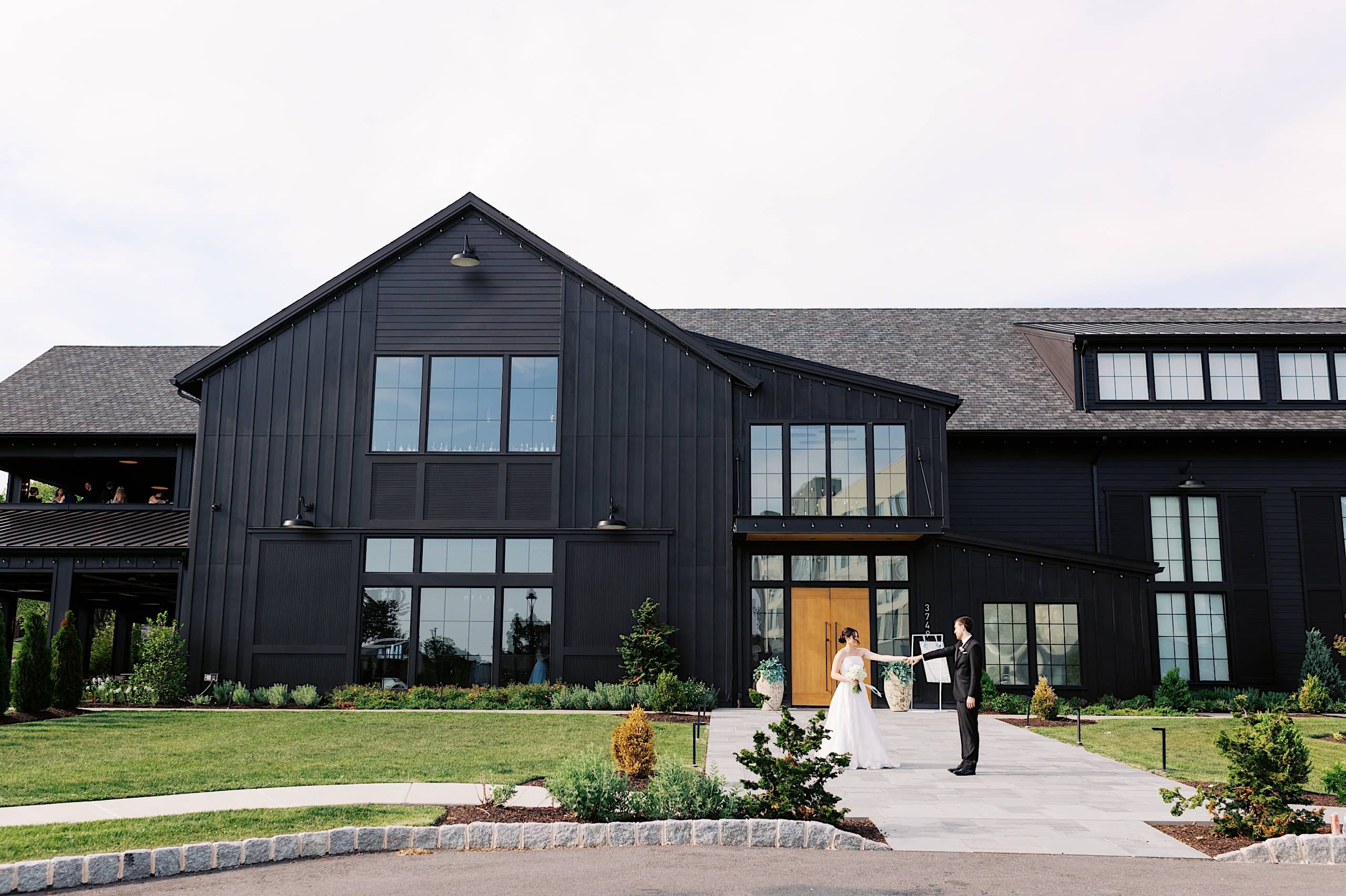 A bride and groom stand outside The Ivy at Ellis Preserve, a large, modern black barn-style building with large windows and landscaped grounds—perfect for a beautiful spring wedding.