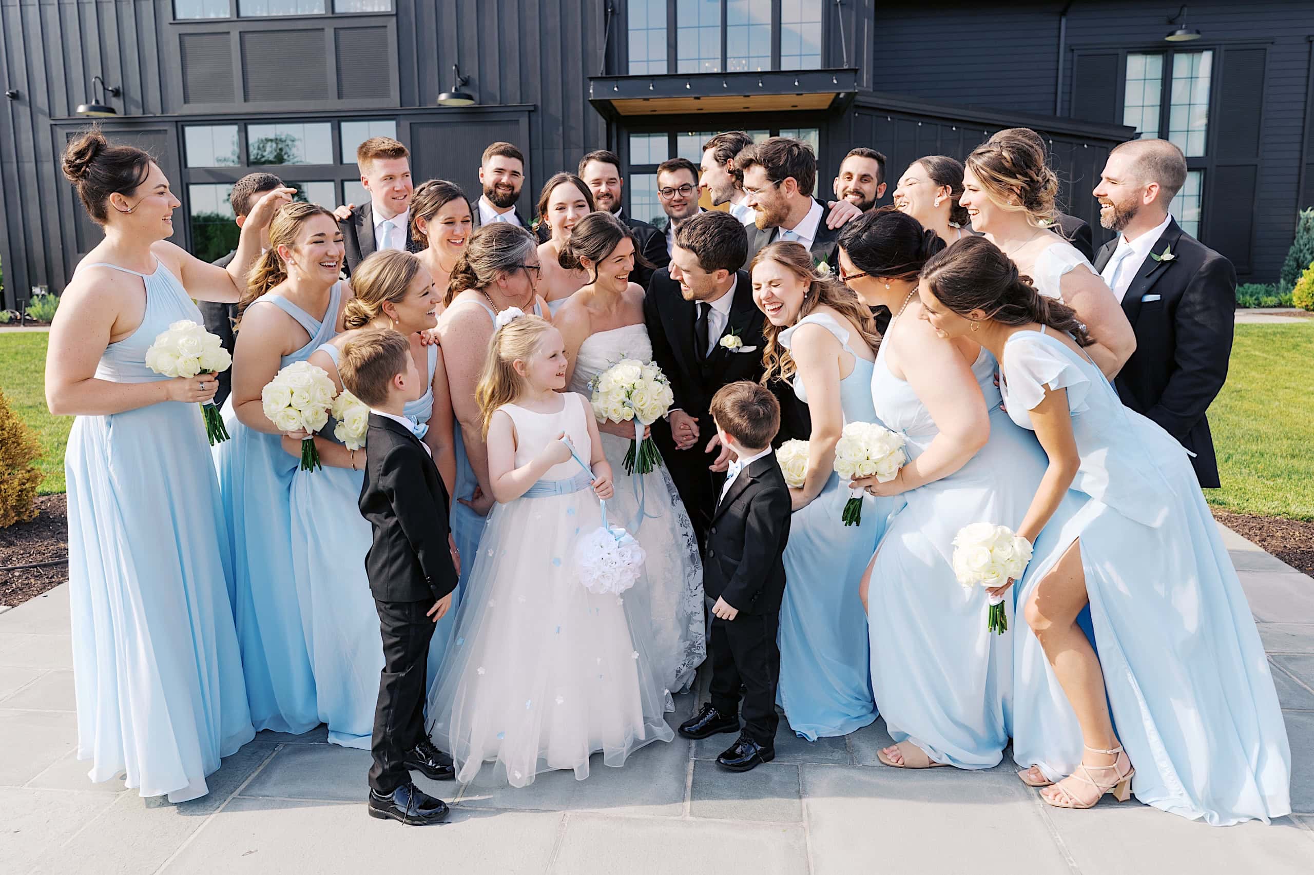 A wedding party poses outdoors at The Ivy at Ellis Preserve spring wedding, with bridesmaids in light blue dresses holding white bouquets and groomsmen in dark suits gathered around the bride and groom. Two children stand in front.