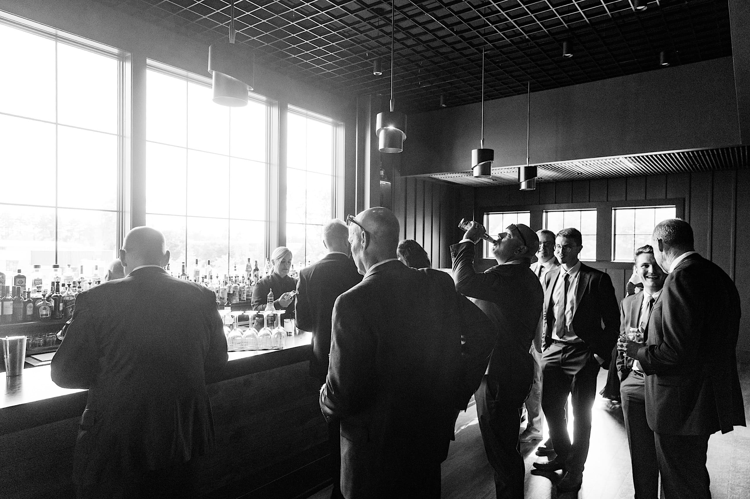 A group of men in suits stand and talk near a bar in a modern, well-lit room with large windows at The Ivy at Ellis Preserve during a spring wedding.
