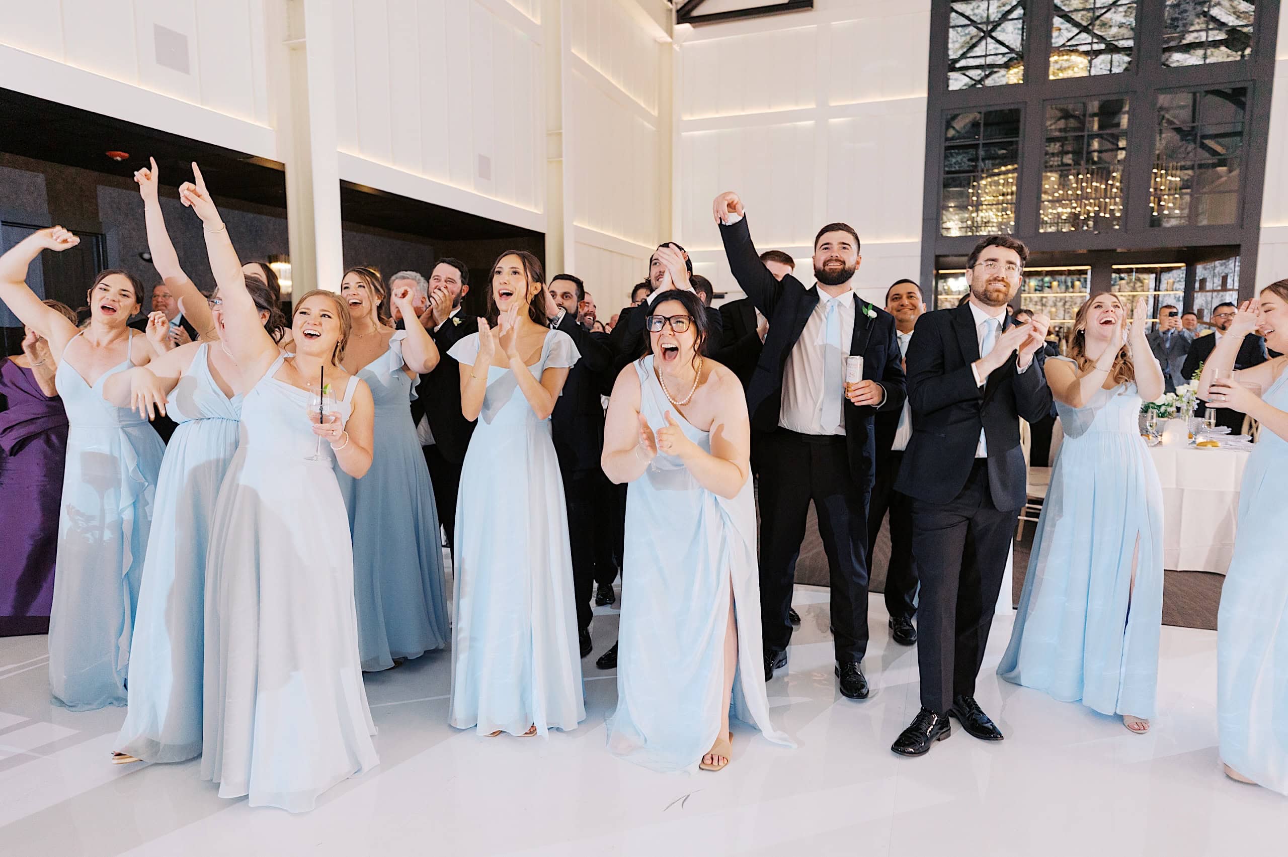 A group of people in formal attire, with women in light blue dresses and men in black suits, stand indoors at The Ivy at Ellis Preserve spring wedding, cheering and pointing, appearing excited and engaged in the event.