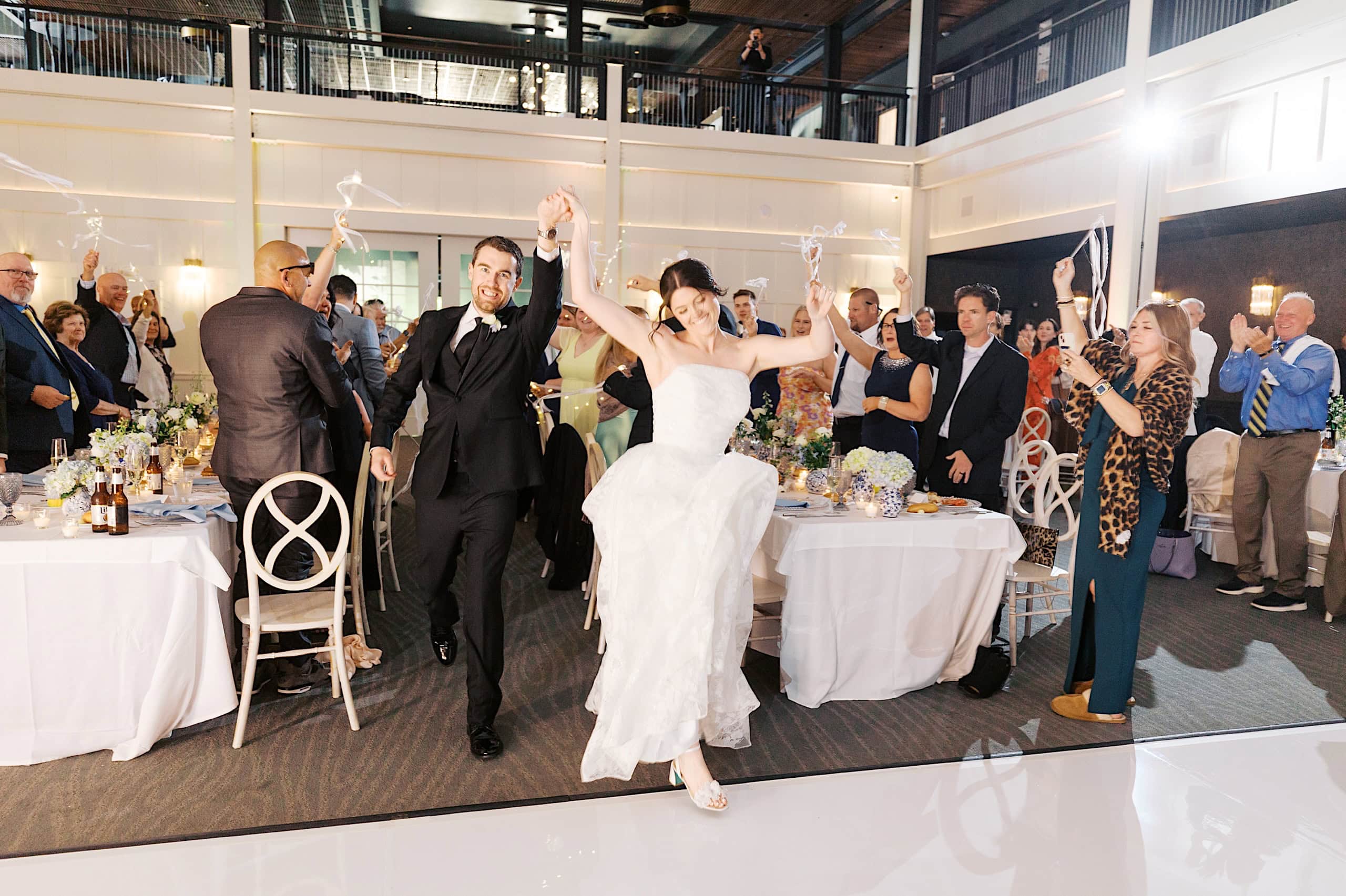 A bride and groom walk hand in hand through their The Ivy at Ellis Preserve spring wedding reception as guests cheer and wave ribbon wands.
